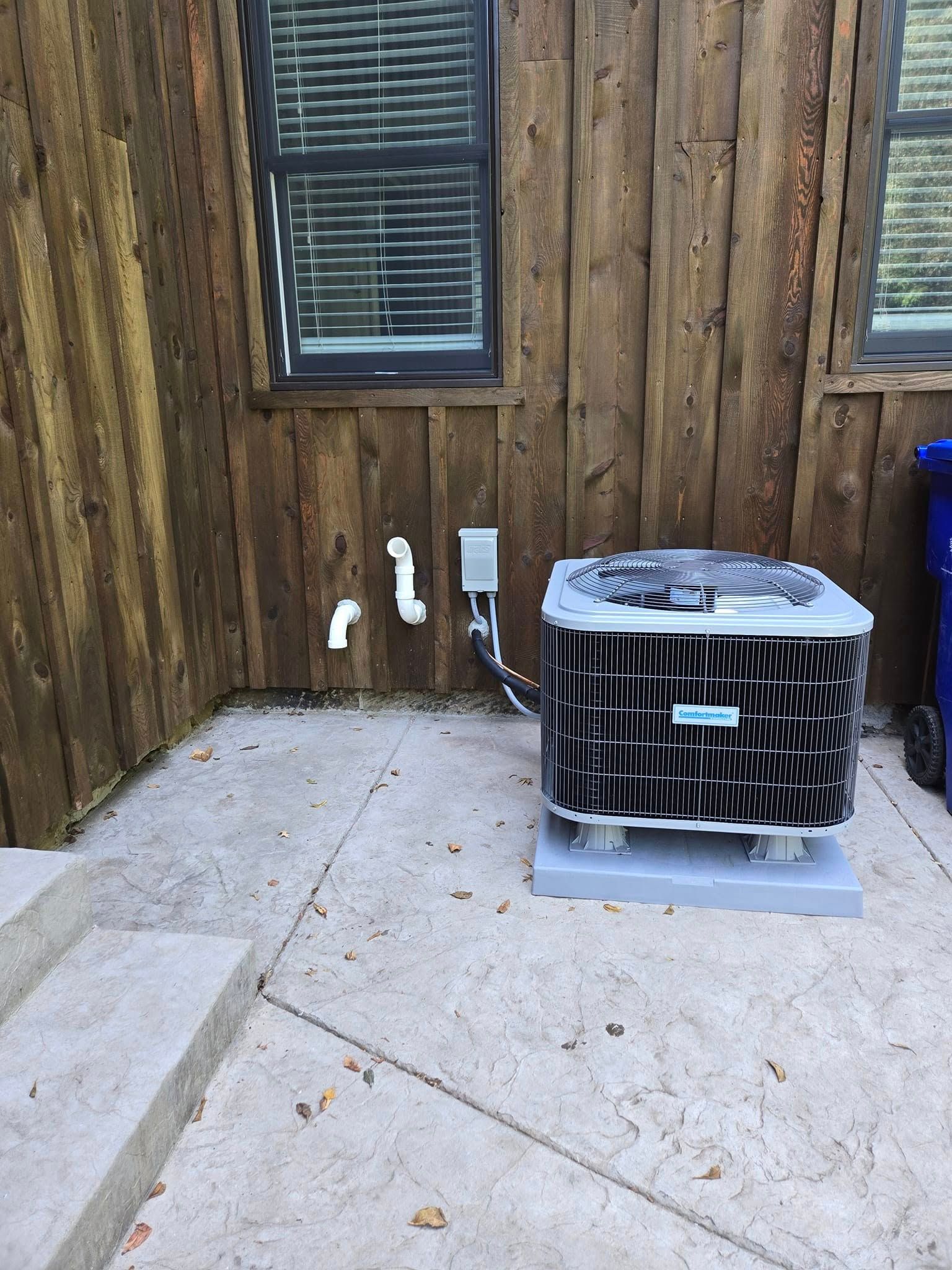 Air conditioning unit outside a wood-paneled building with window and nearby plumbing on a concrete slab.