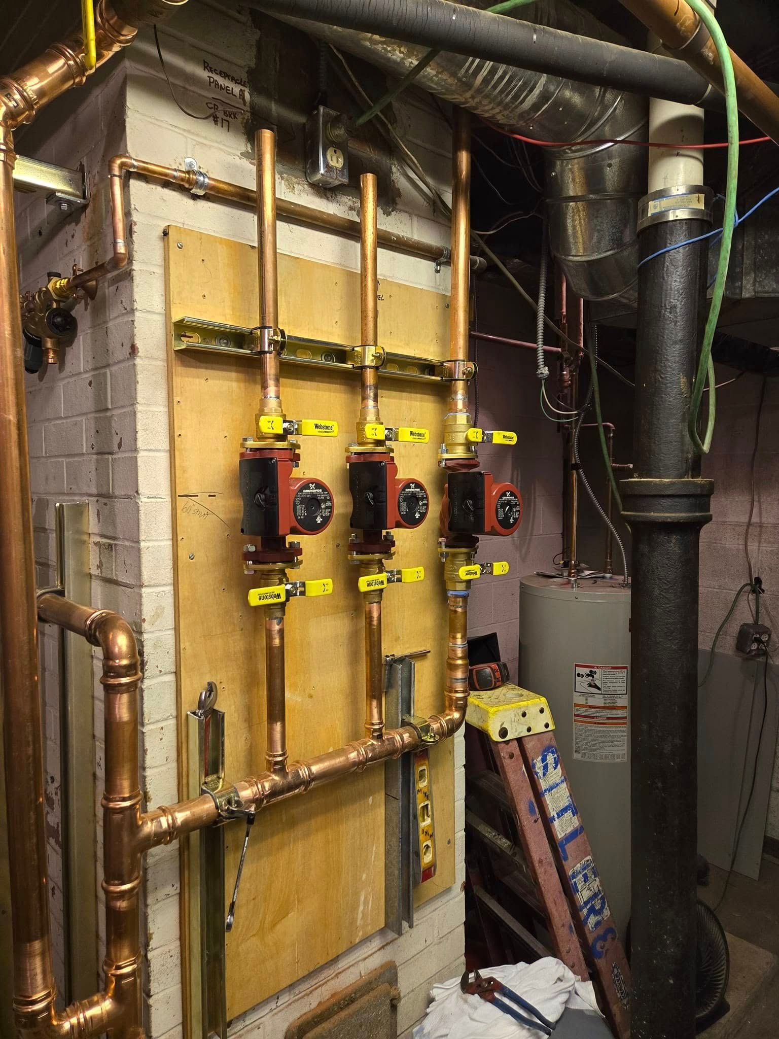 Copper pipes and pumps on a wooden panel in a utility room. Boiler tank visible in the background.