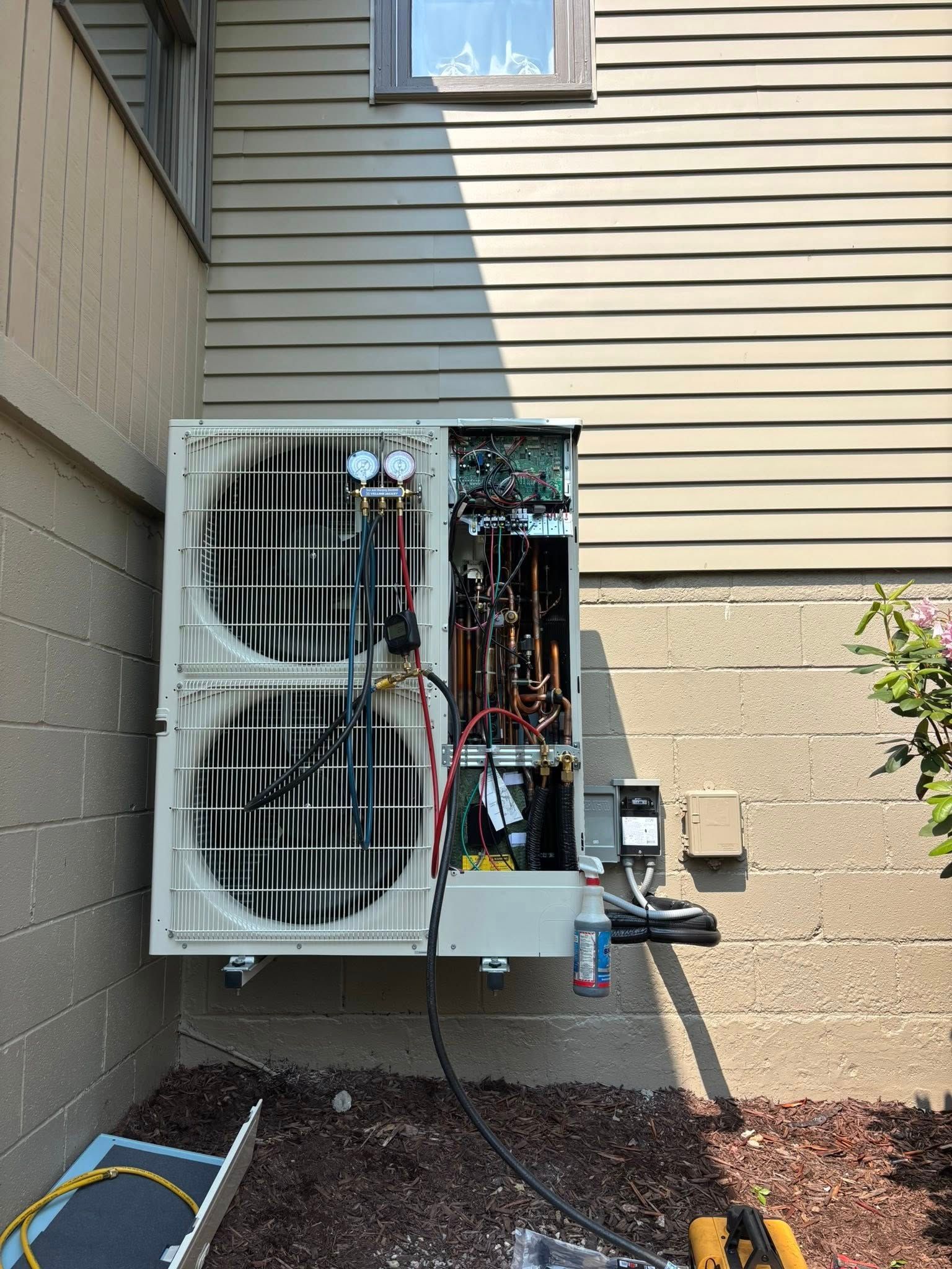 Opened outdoor HVAC unit mounted on a beige wall, next to a window. Wires and components are visible inside.