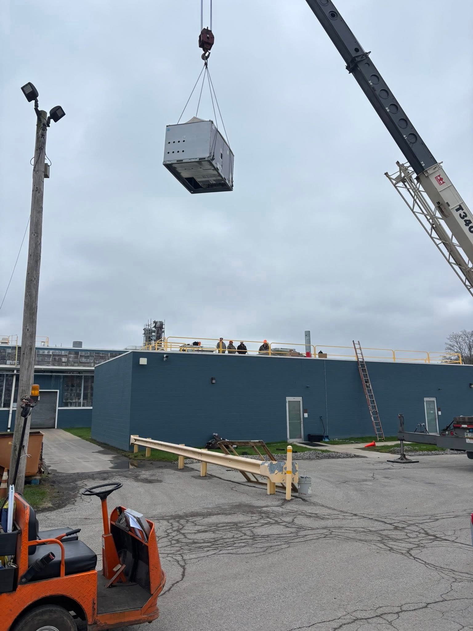 A crane lifts a large, gray unit above a dark blue industrial building.  An orange forklift sits nearby.