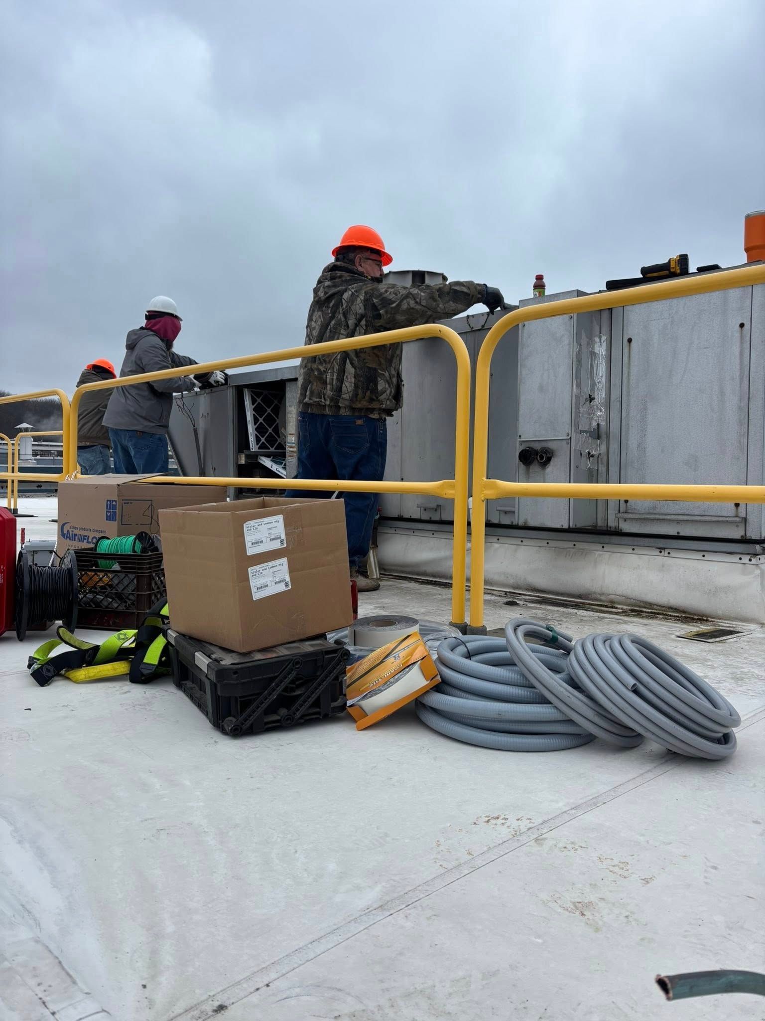 Workers in hard hats on a rooftop near equipment, with tools and supplies. Cloudy sky.