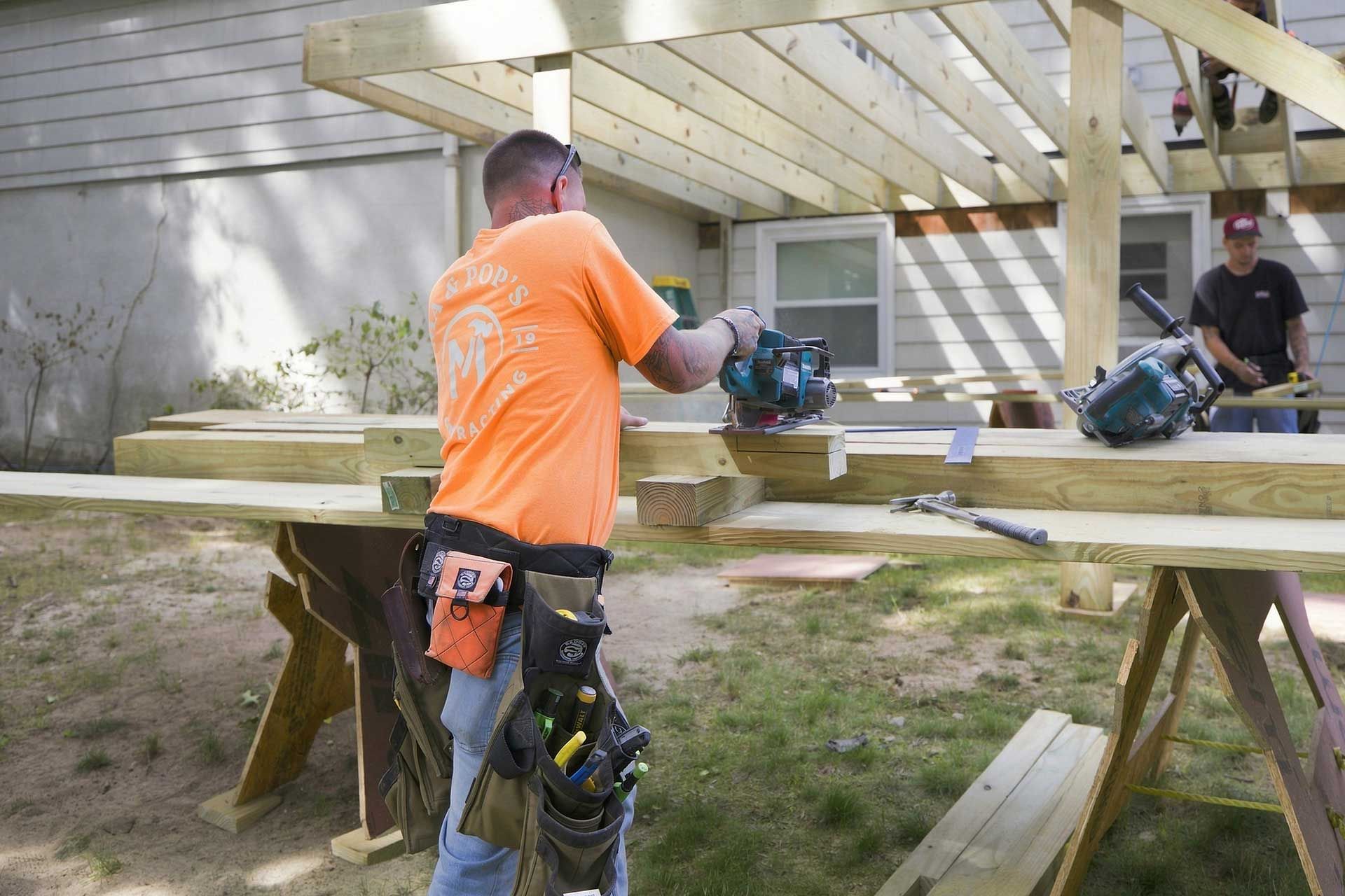 Two workers cutting wooden boards on sawhorses outdoors, one in an orange shirt and tool belt.
