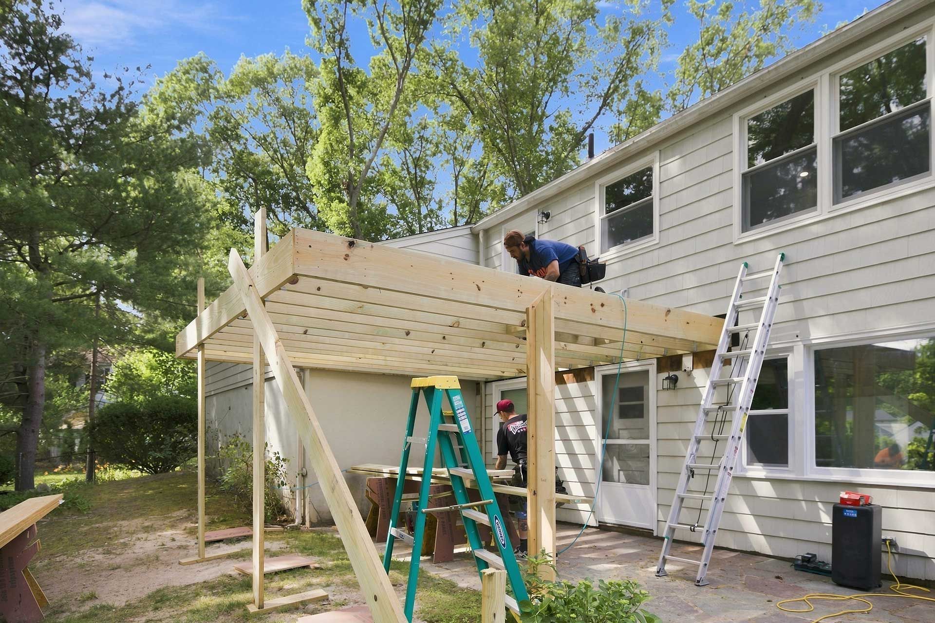 Two workers building a raised wooden deck beside a house, with ladders and tools in the yard