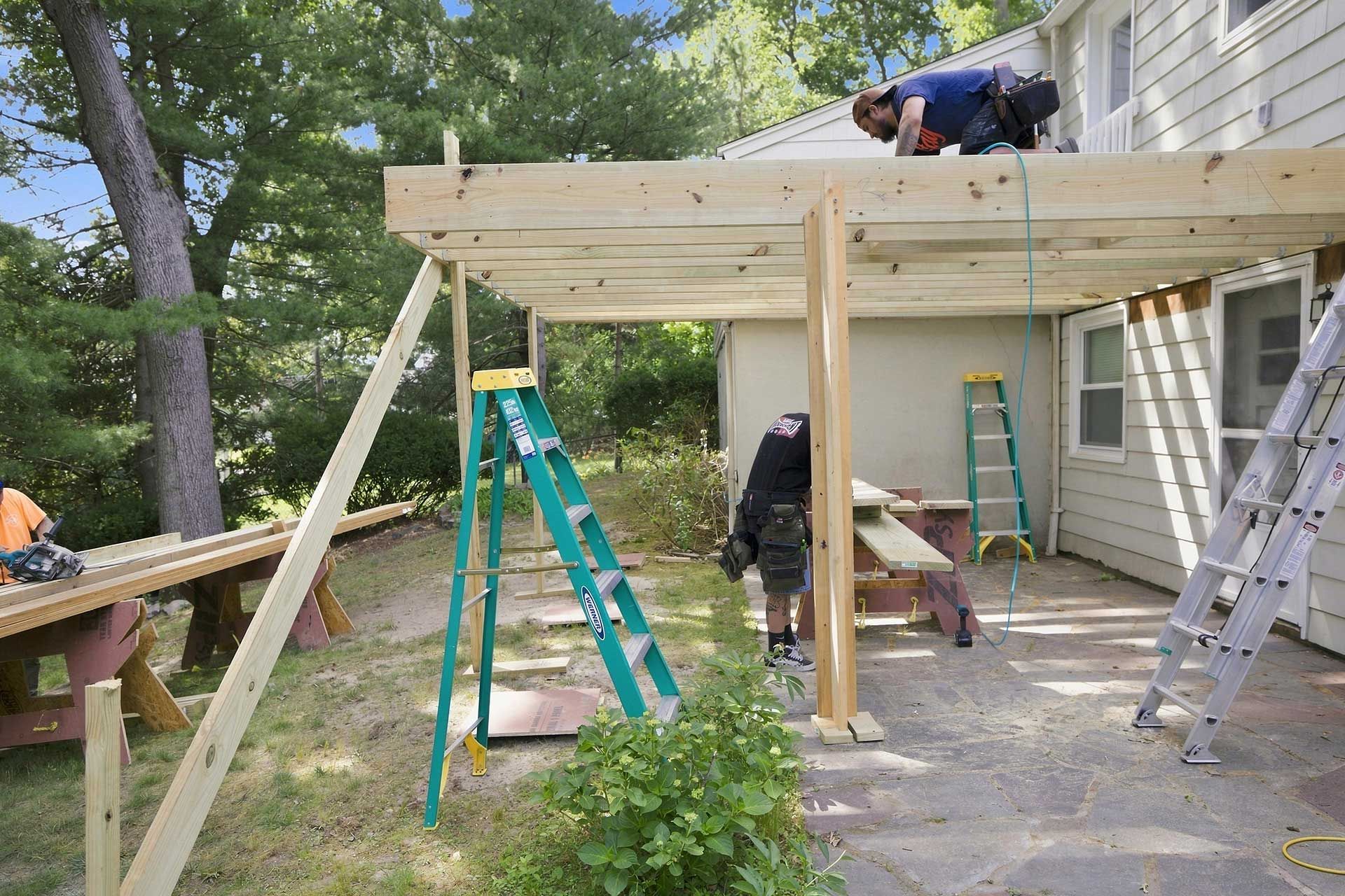 People building a wooden deck beside a house, with ladders and tools on a driveway.