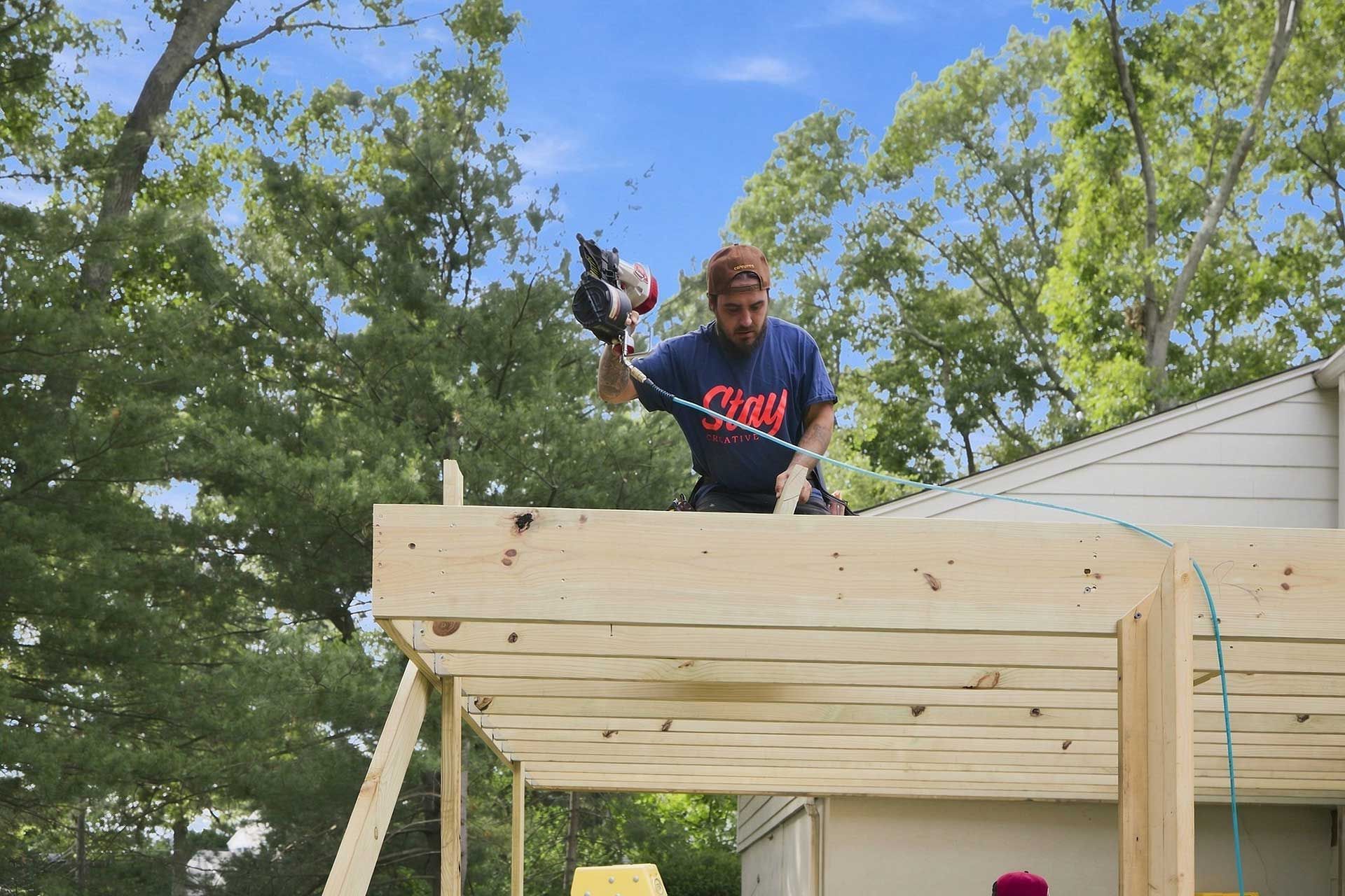 Person using a power drill on a wooden deck frame outdoors beside a house.
