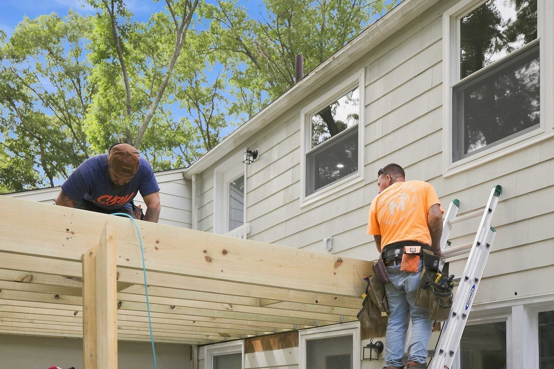 Two workers install a large wooden deck beam beside a house, one on a ladder and one leaning over the beam.