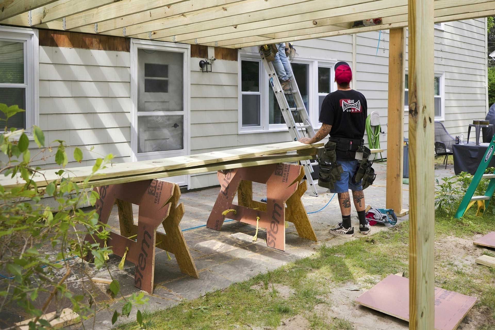 Worker beneath a house deck, inspecting a window area with tools and sawhorses nearby