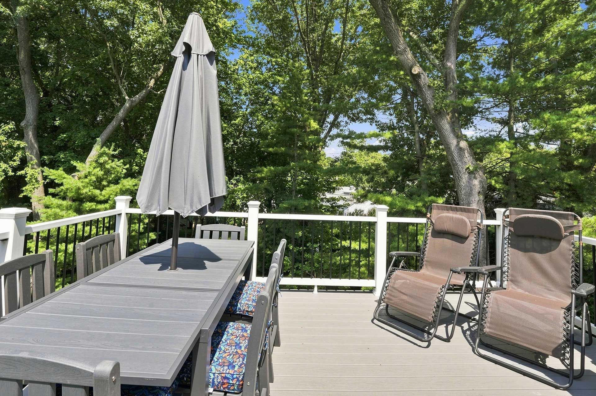 Deck with patio table, closed umbrella, and two lounge chairs overlooking trees and a wooded yard