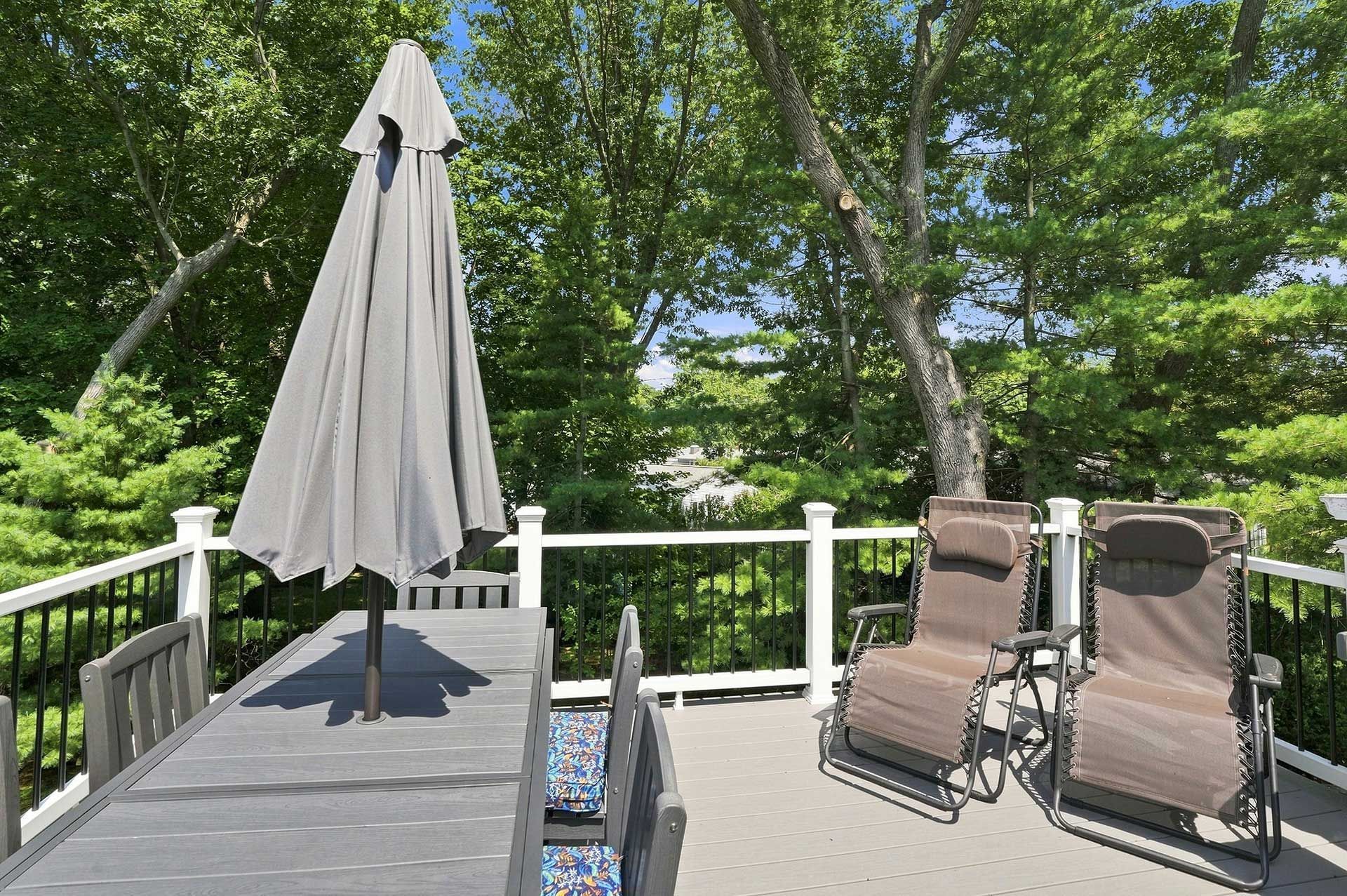 Patio deck with folded umbrella, two lounge chairs, and green trees in the background