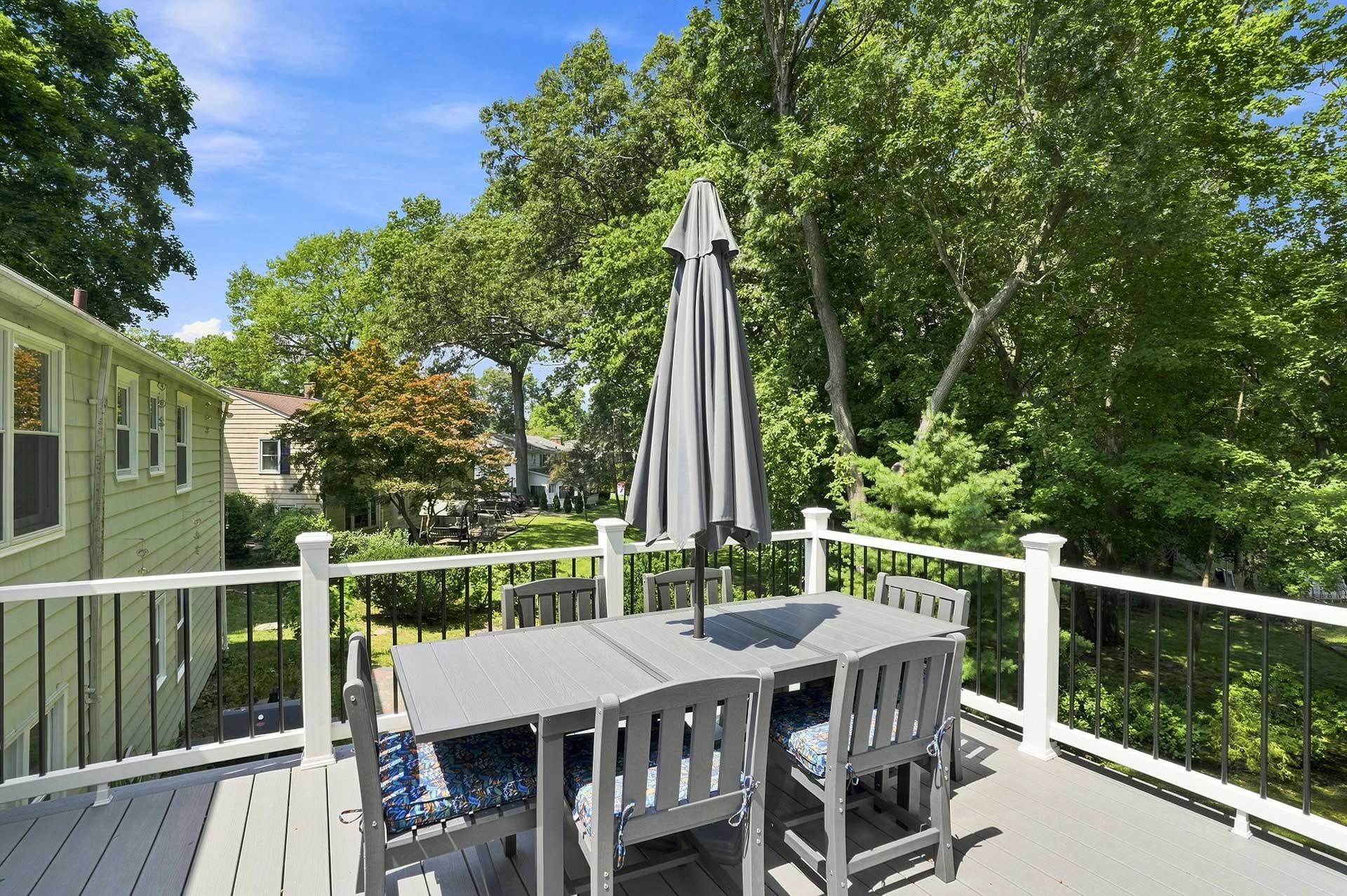 Wooden deck with outdoor table, chairs, and umbrella, overlooking green trees and a neighboring house