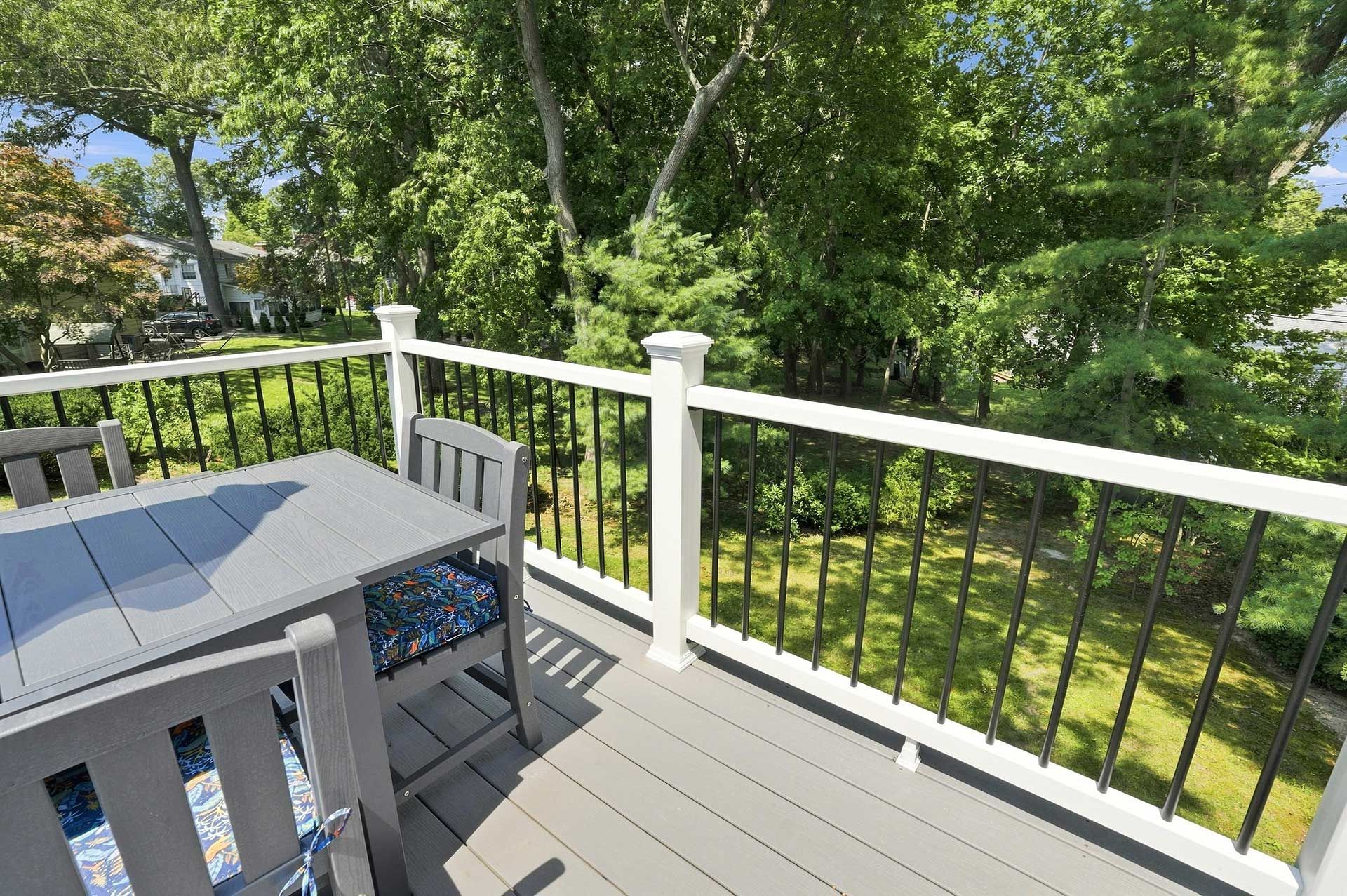 Sunlit deck with gray table and chairs, white railing, and green trees beyond.