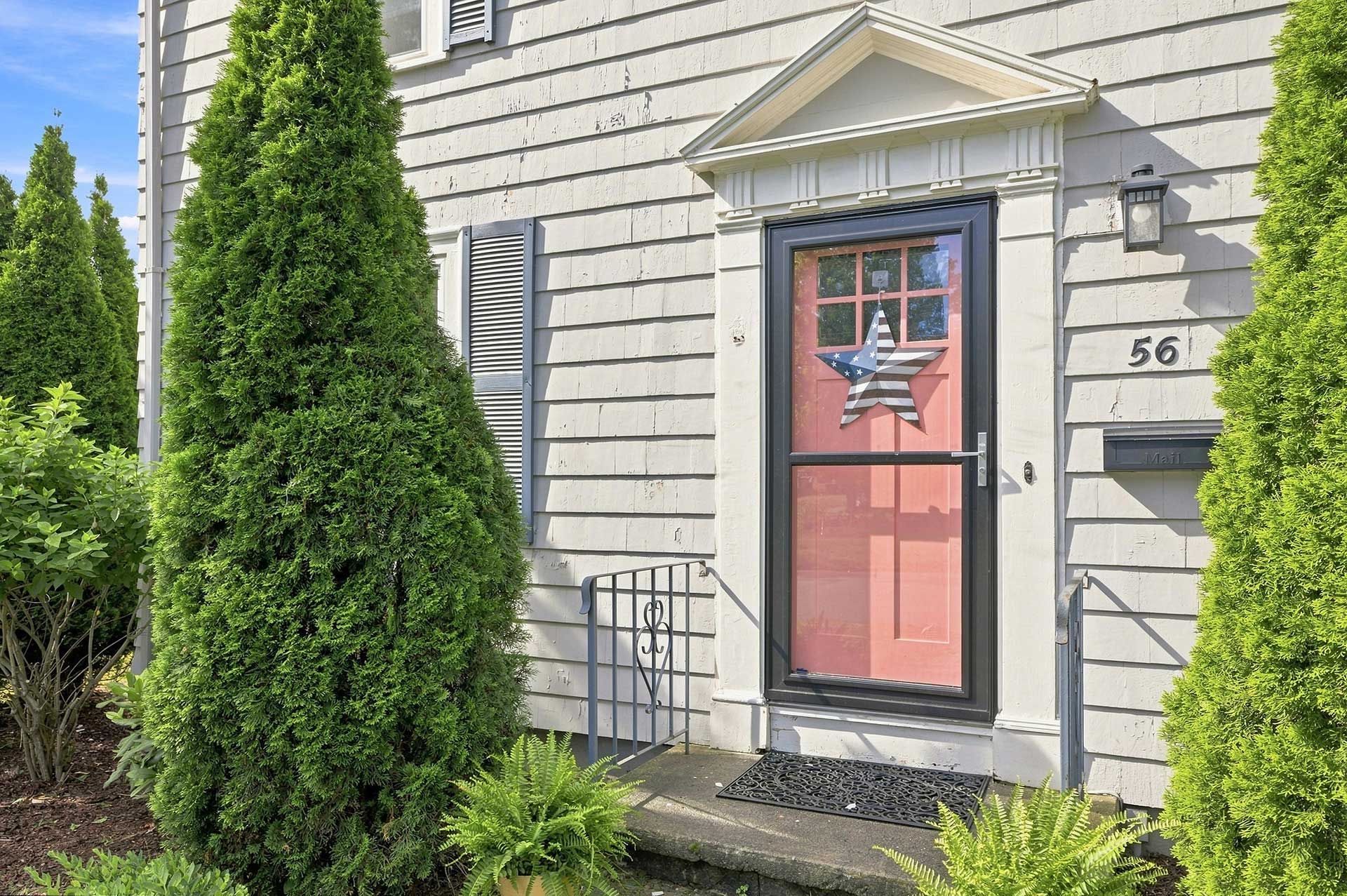Pink front door with American flag wreath on a  house, framed by tall evergreen shrubs