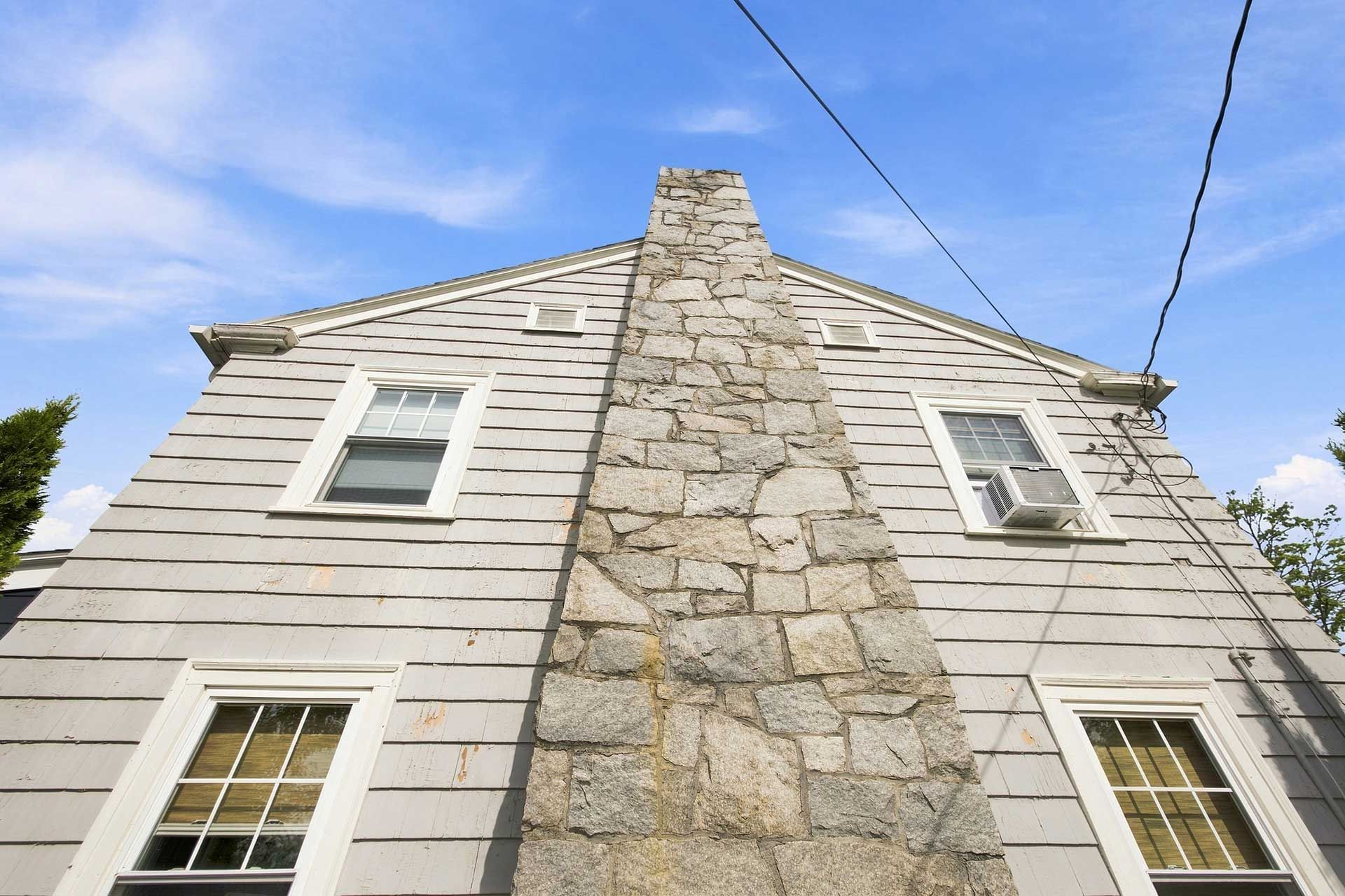 Stone chimney on a white house with two windows, viewed from below against a blue sky.