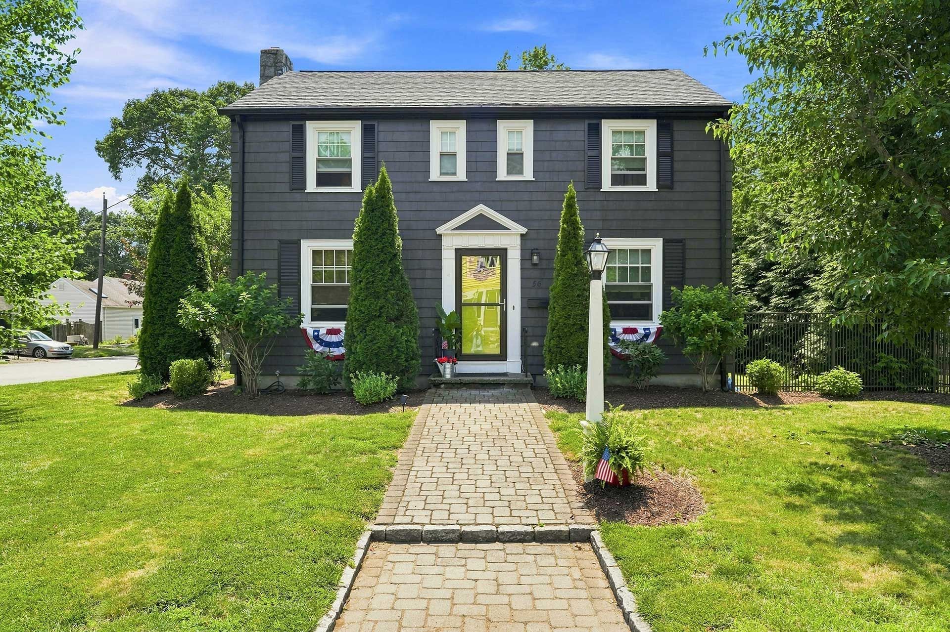 Dark gray house with lime green door and brick walkway, framed by tall evergreens and a green lawn.
