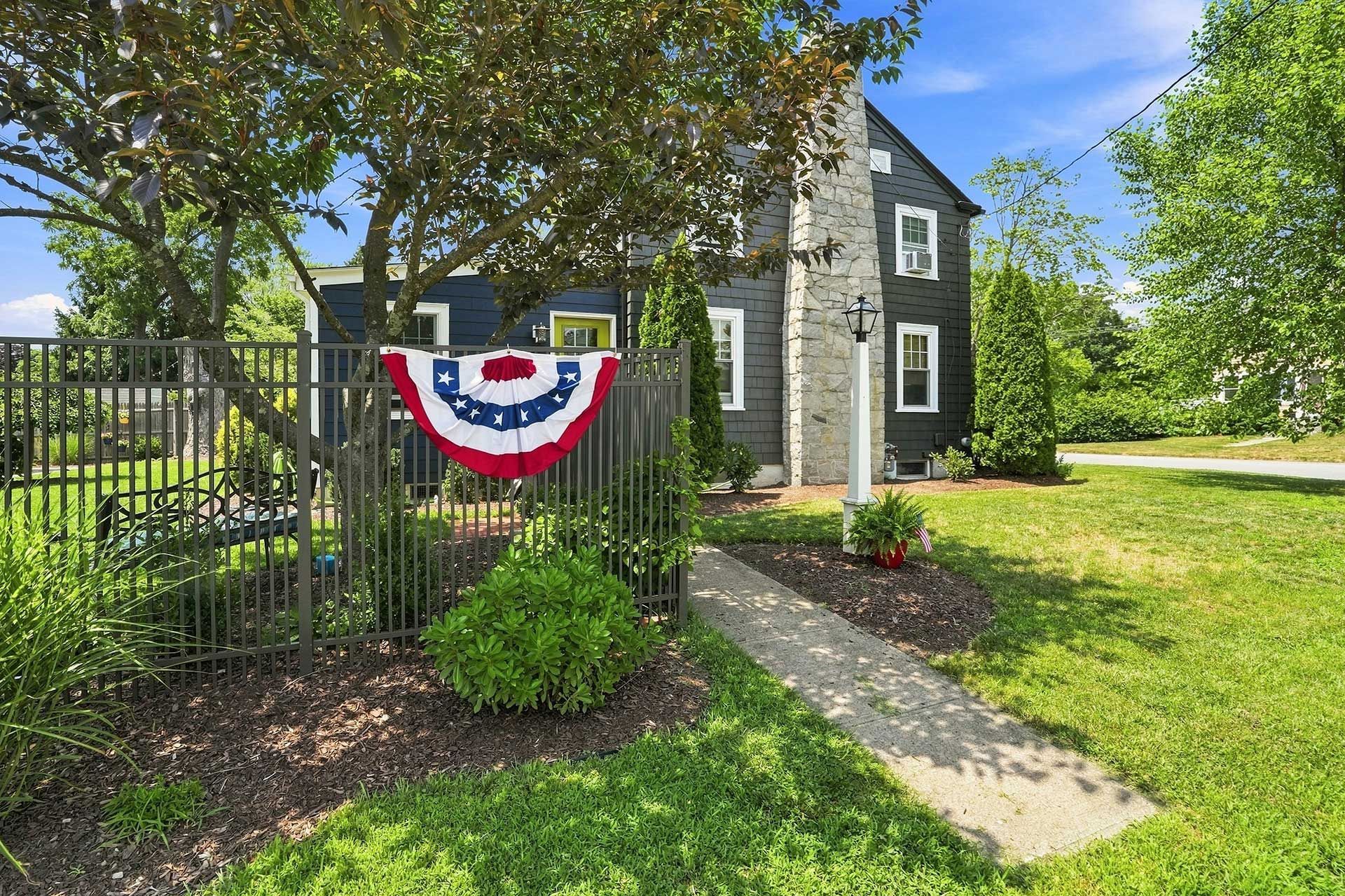 Dark gray house with white trim and patriotic bunting by a garden path, beside a black fence and lawn.