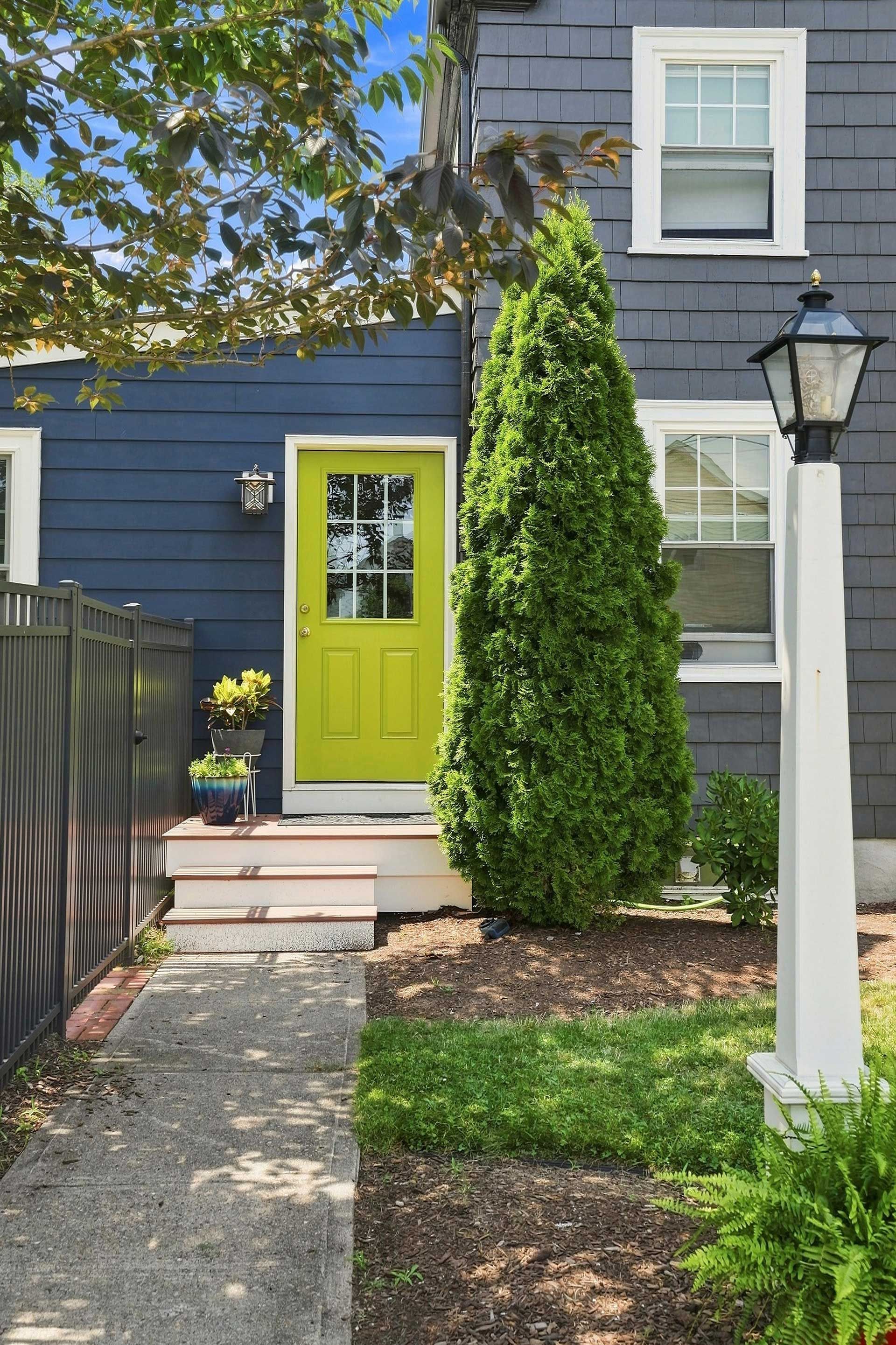 Blue house exterior with lime-green front door, stone path, and tall evergreen shrub by the entrance