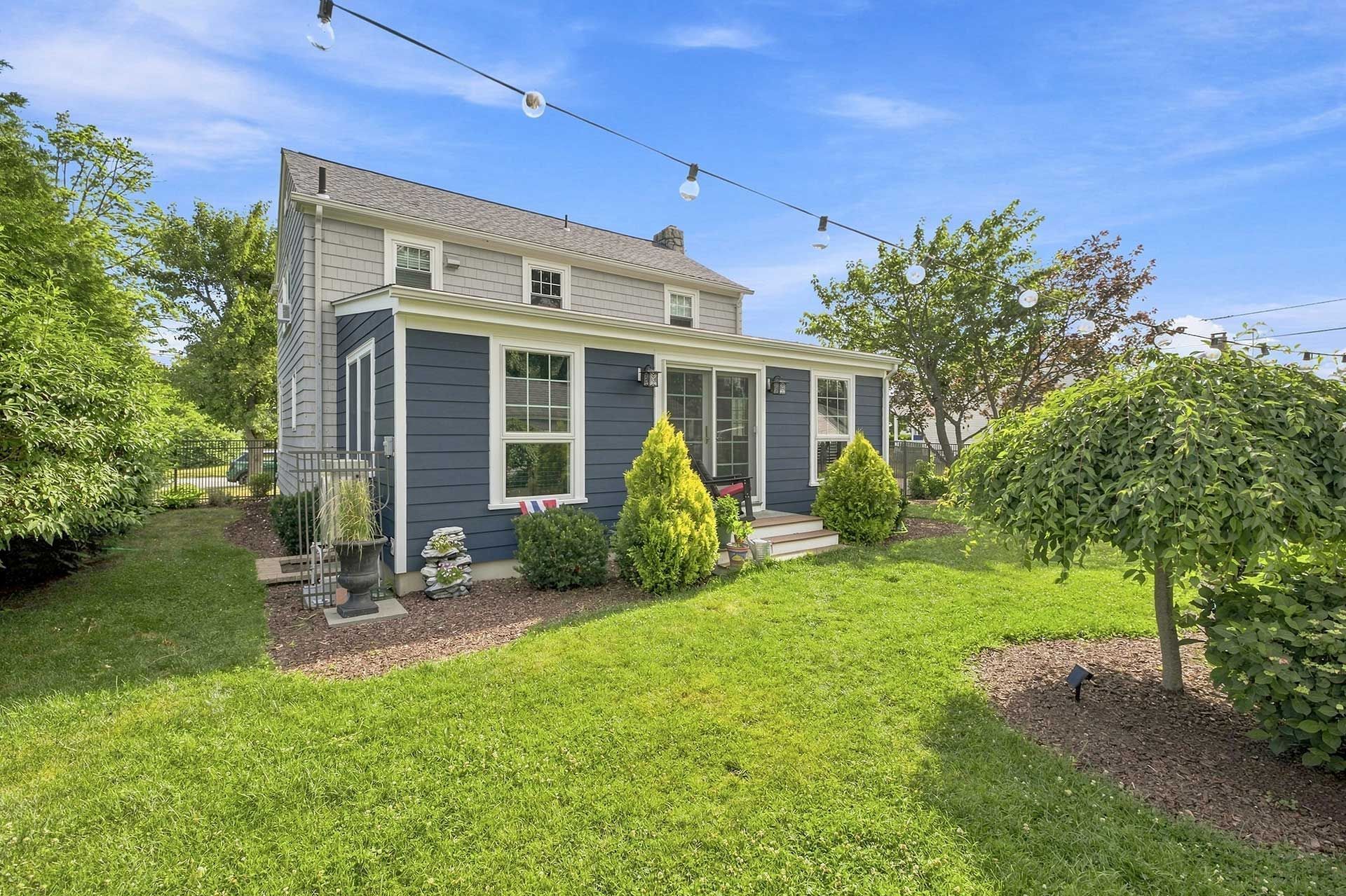 Blue-gray house with white trim, a green lawn, and string lights in a sunny yard.