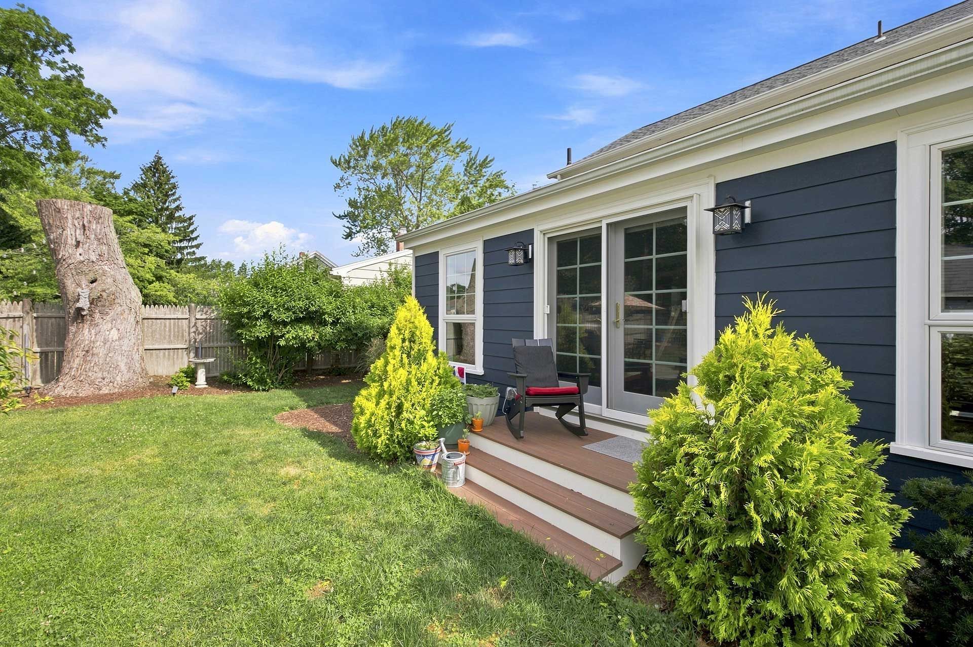 Backyard patio with dark blue house, glass doors, steps, and small shrubs beside a grassy lawn.