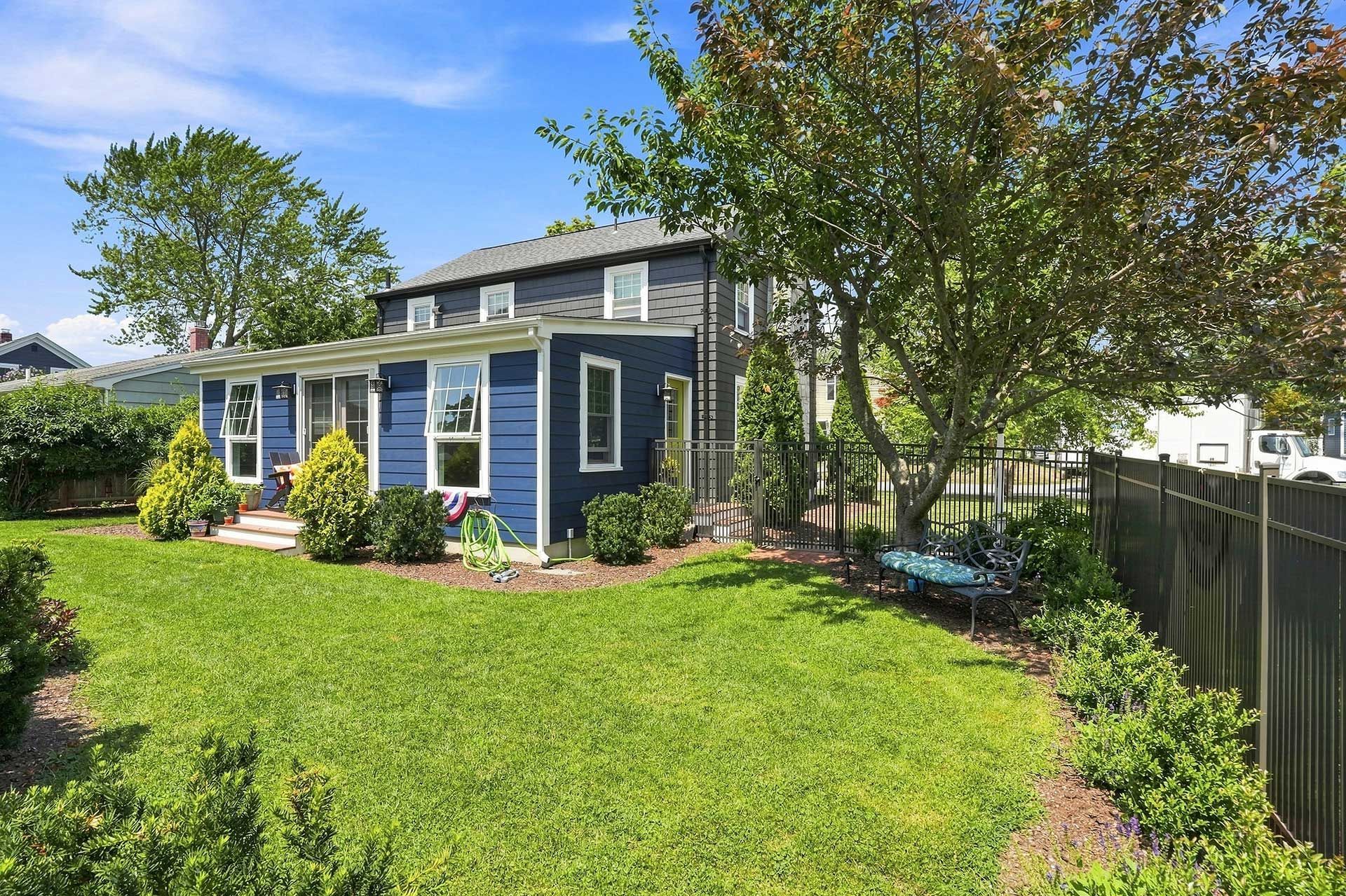 Blue house with white trim and a green lawn beside a fenced backyard and trees.