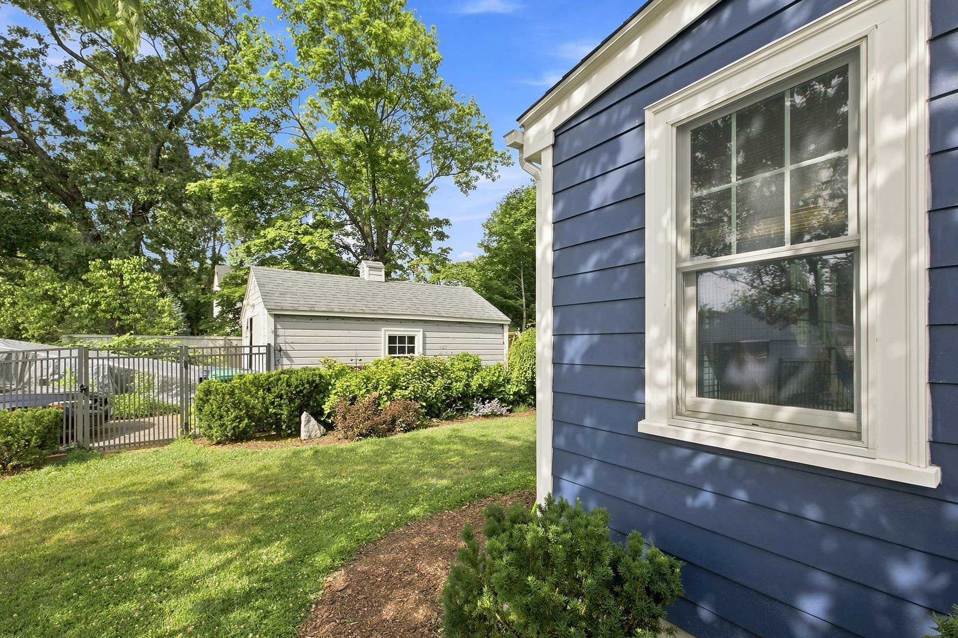 Blue house exterior with white-trimmed window, green lawn, and fenced backyard under sunny trees