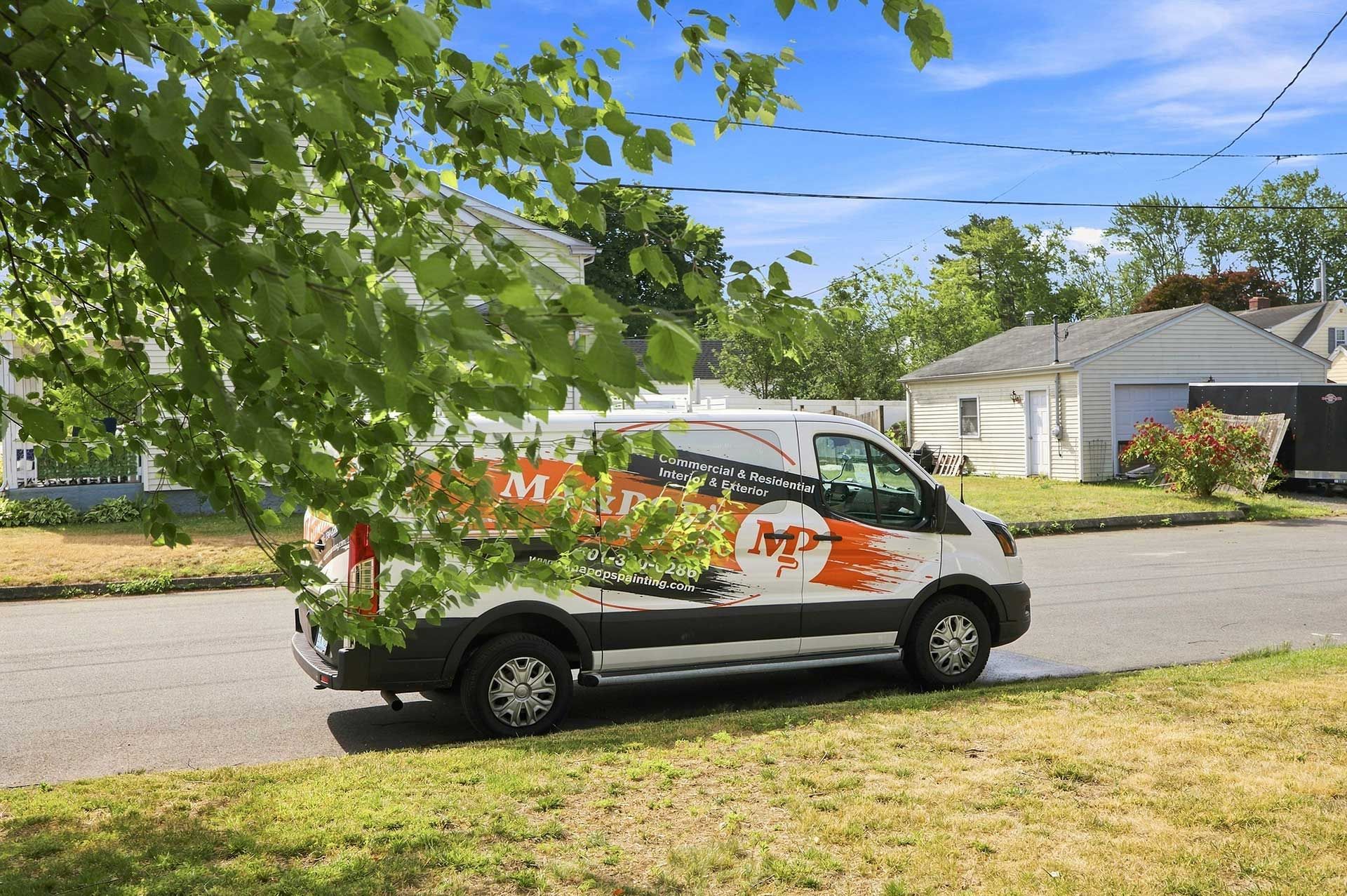 White van with graphics parked on a suburban street, viewed through leafy branches