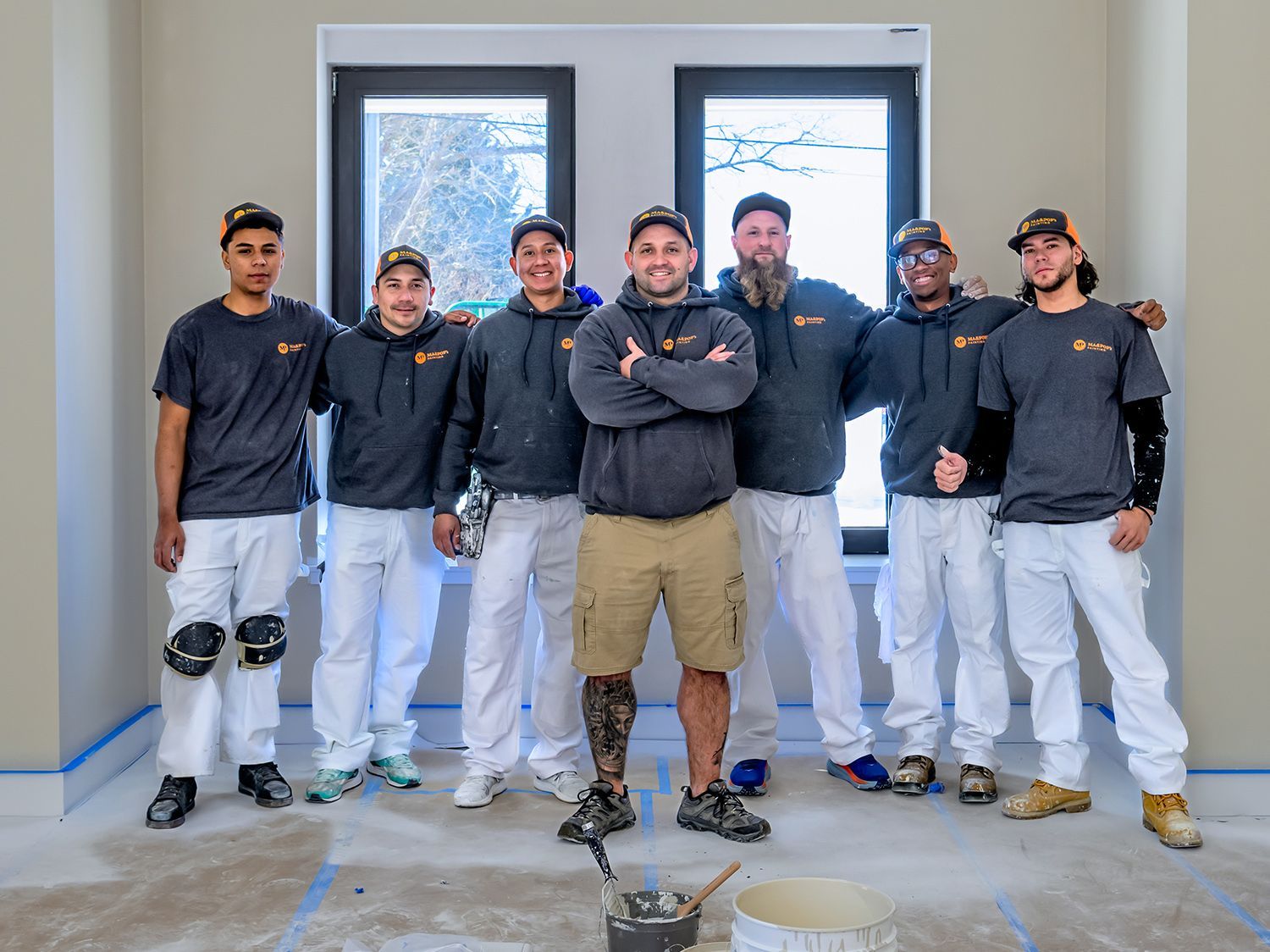 Painting crew posing in a room; the leader has crossed arms. They wear gray shirts, white pants, and kneepads.