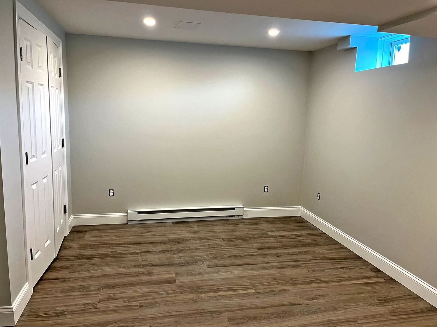 Empty, neutral-toned room with white doors, baseboards, and window, a heating element, and wood-look flooring.