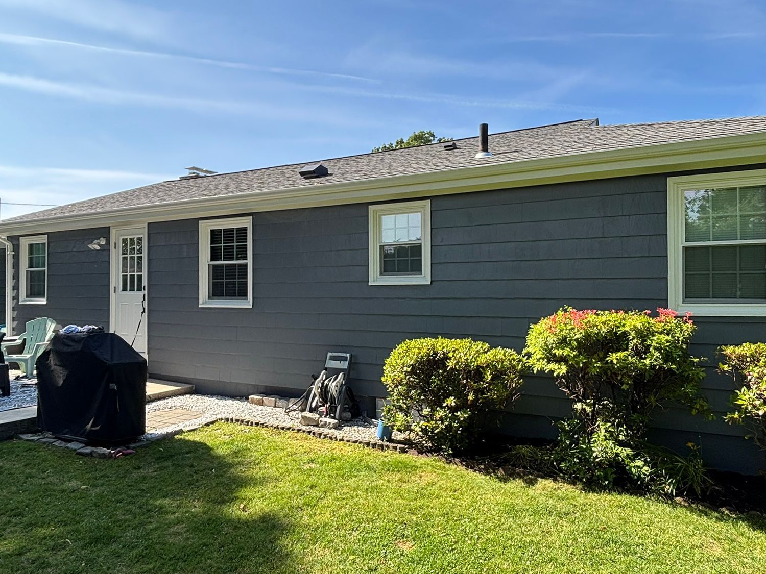 Gray-sided house with white trim, windows, and door. A covered grill, bushes, and a green lawn are in the yard.