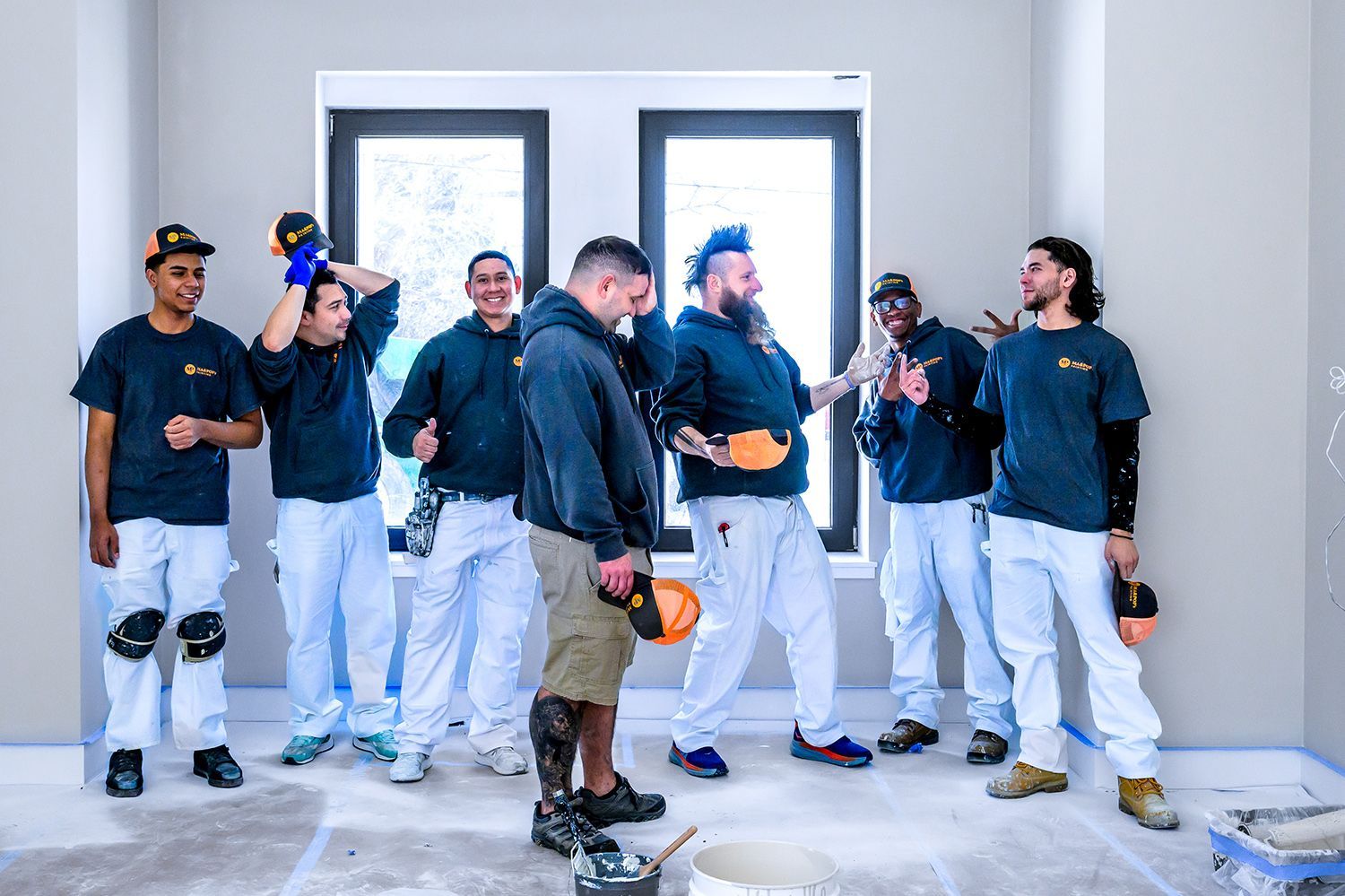Group of painters in a room, some smiling, wearing paint-covered pants and black shirts.