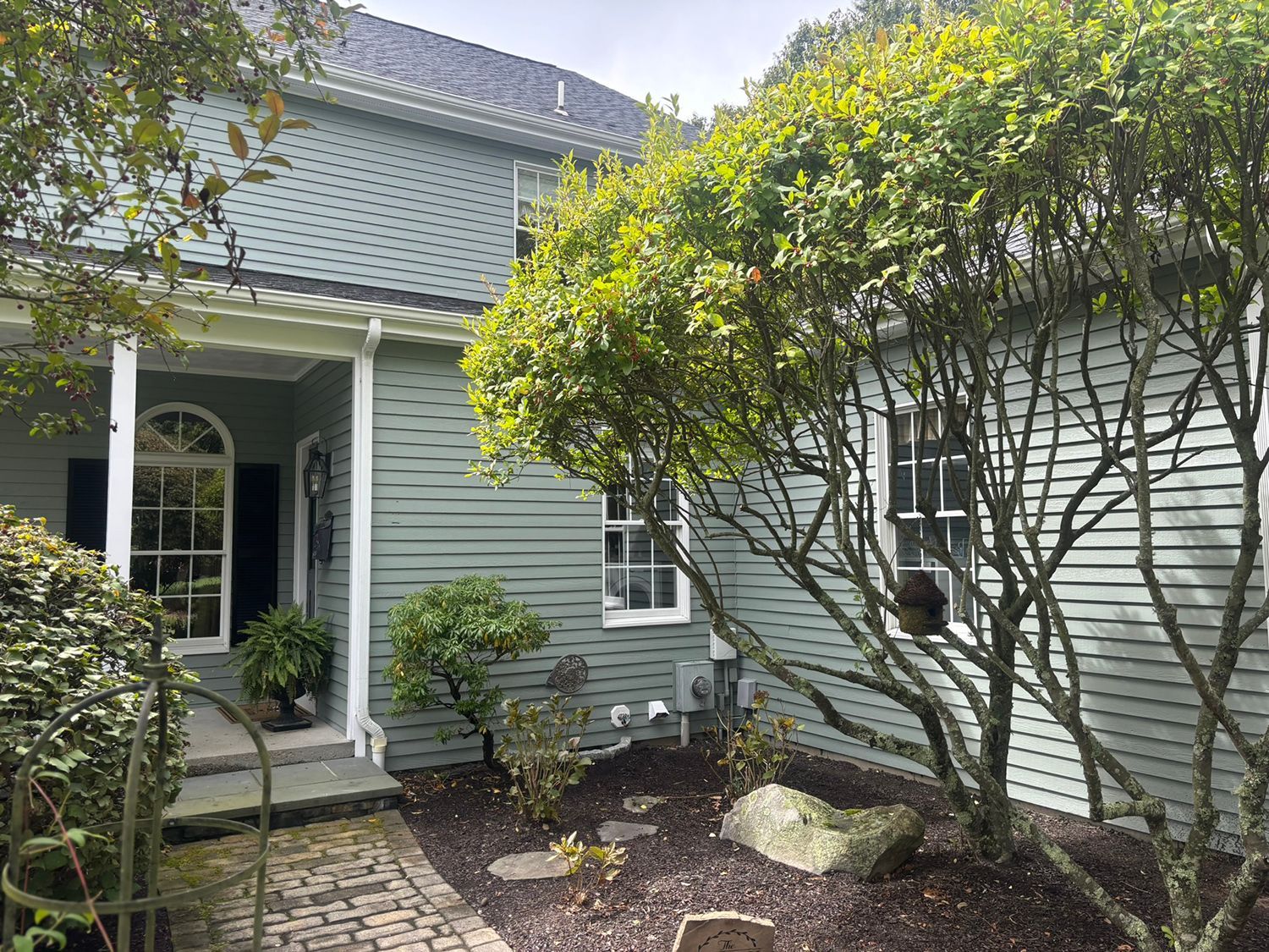A light green house with a brick walkway, trees, and a partially visible roof.