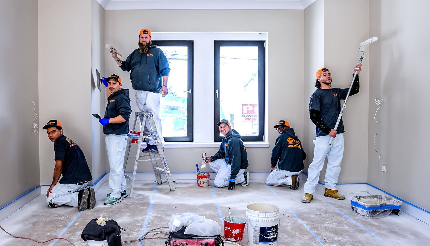 Six people paint a room. Some are on ladders or kneeling. Walls are beige, with blue painter's tape on the floor.