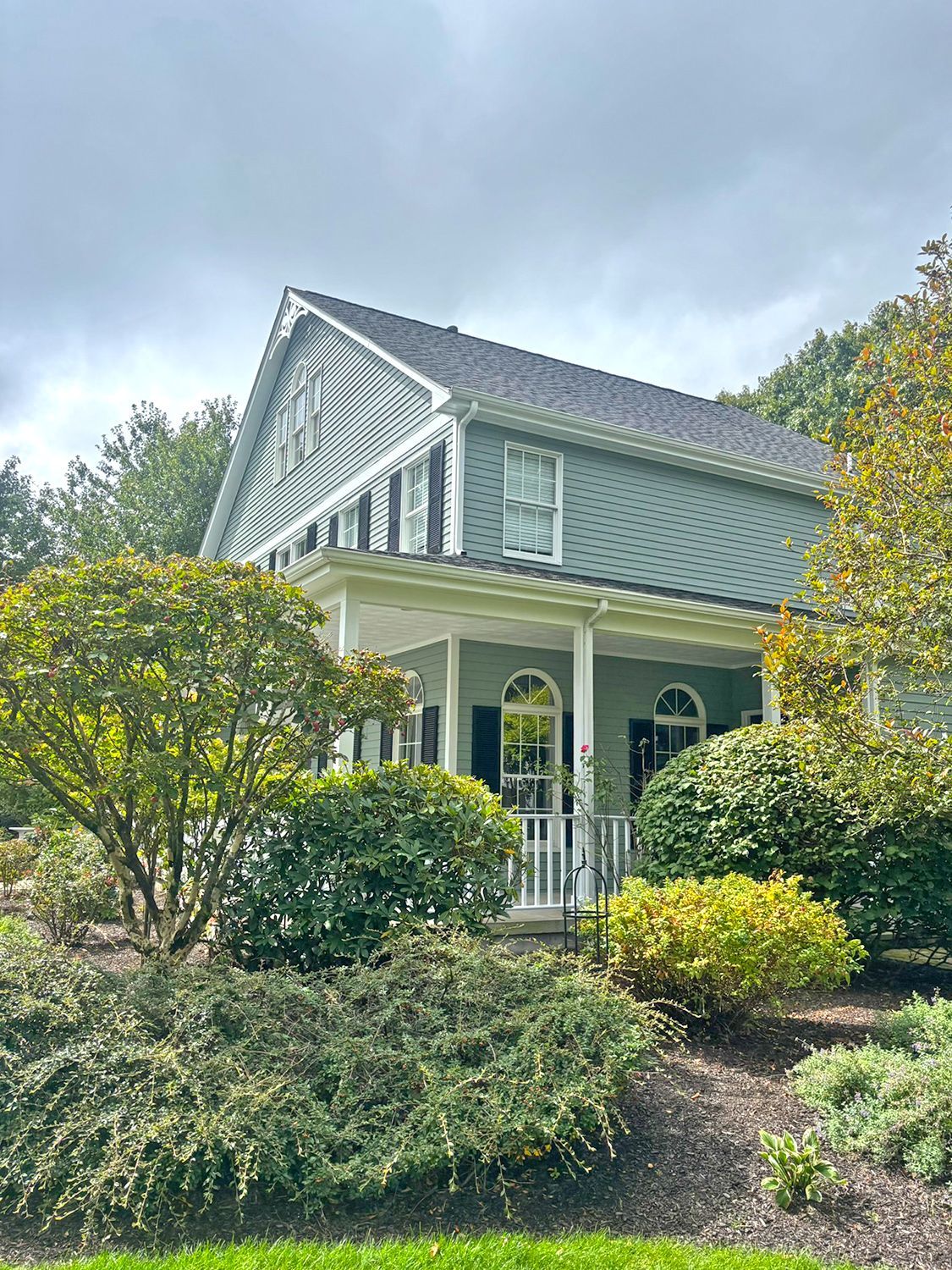 Two-story blue-green house with white porch and black shutters, surrounded by green shrubs and trees, overcast sky.
