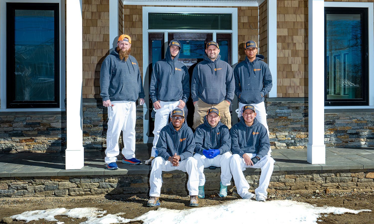 Group of workers in gray hoodies and white pants pose in front of a house.