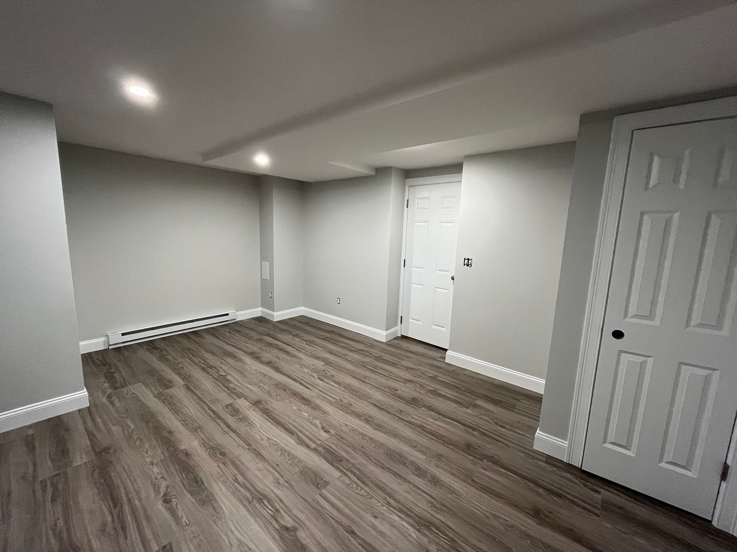 Empty, gray-walled room with wood-look flooring and a white door. Recessed lighting.