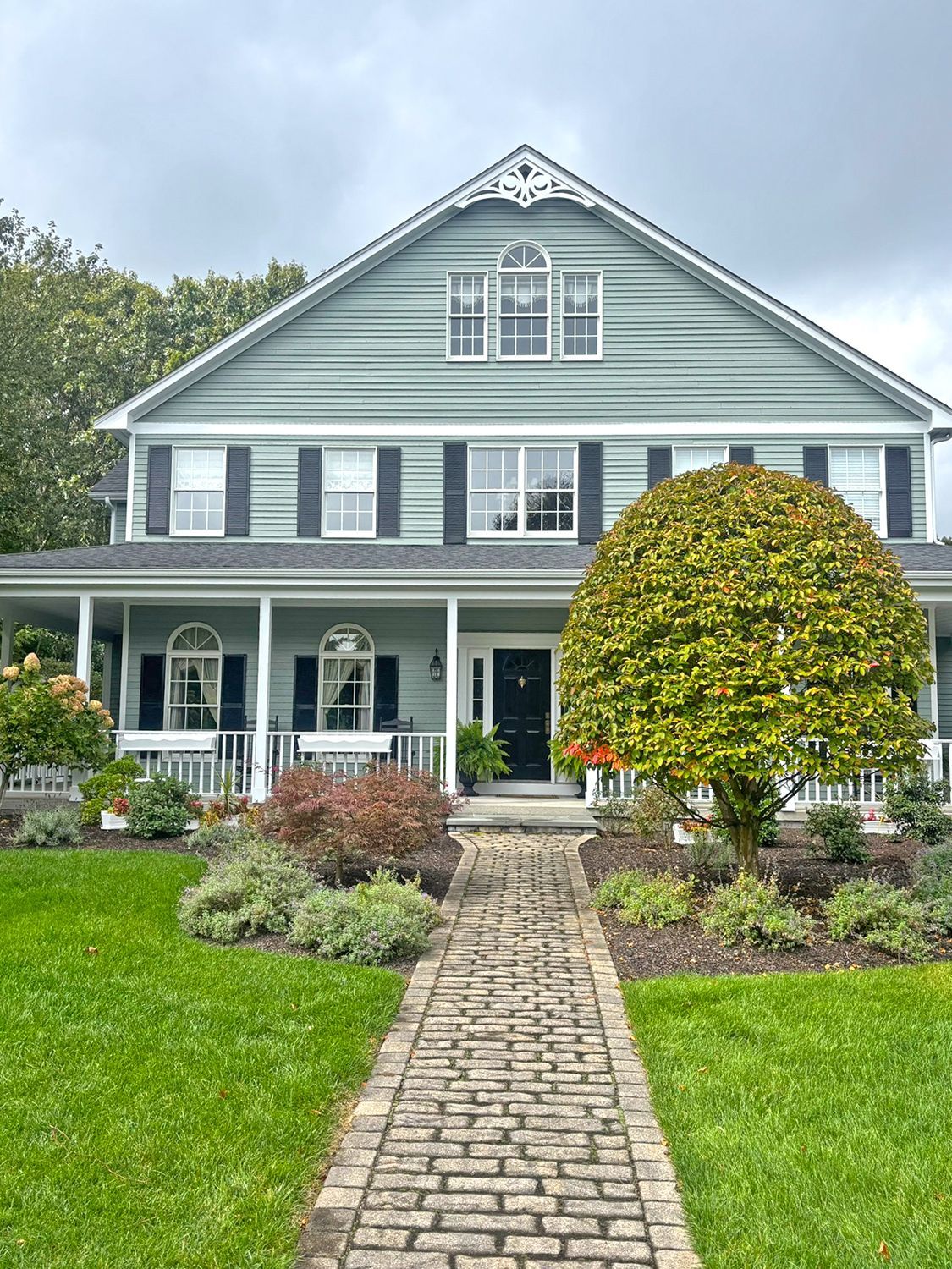 Two-story house with green siding, black shutters, and a porch. A brick path leads to the front door.