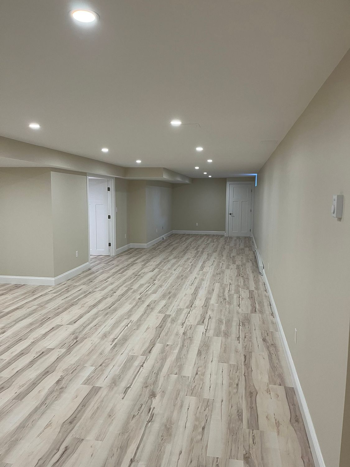 Empty, finished basement with beige walls, light wood-look flooring, recessed lights, and two white doors.