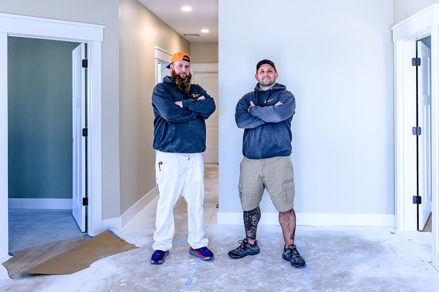 Two men in a room under construction, arms crossed. One has a beard, paint-covered pants, and a cap. The other has a prosthetic leg.