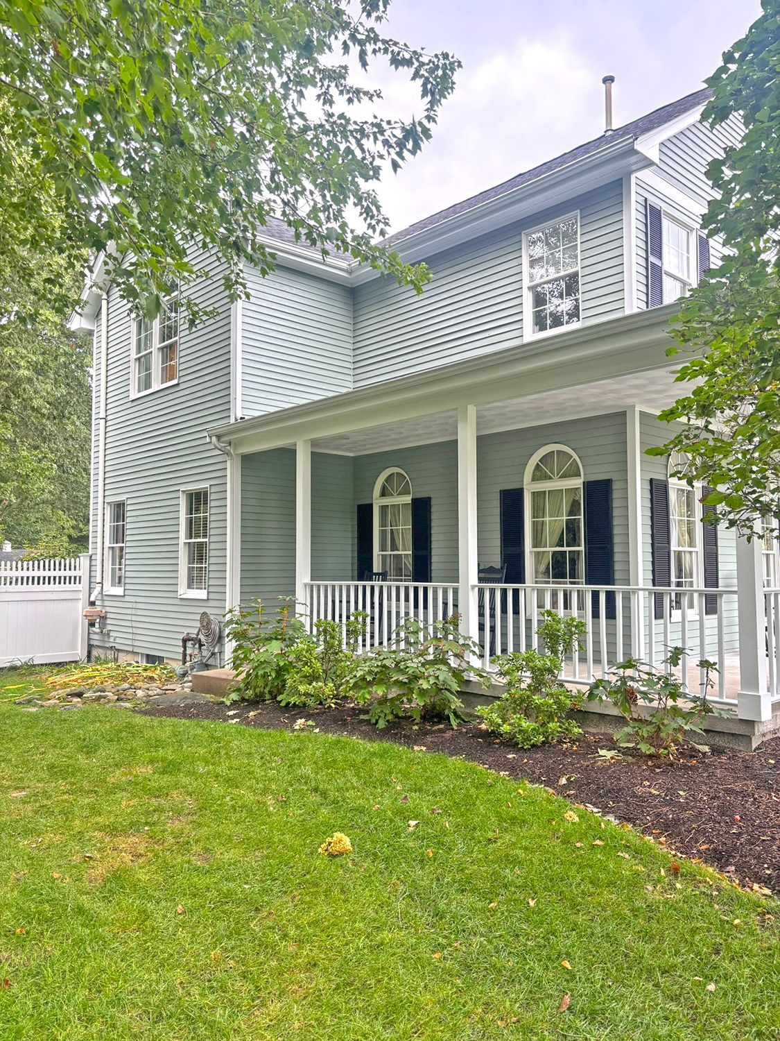 Two-story house with green siding, a white porch, and black shutters, set in a grassy yard.