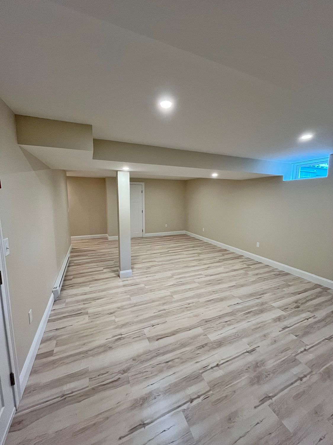 Empty basement with light wood-look flooring, beige walls, recessed lights, and a support beam.