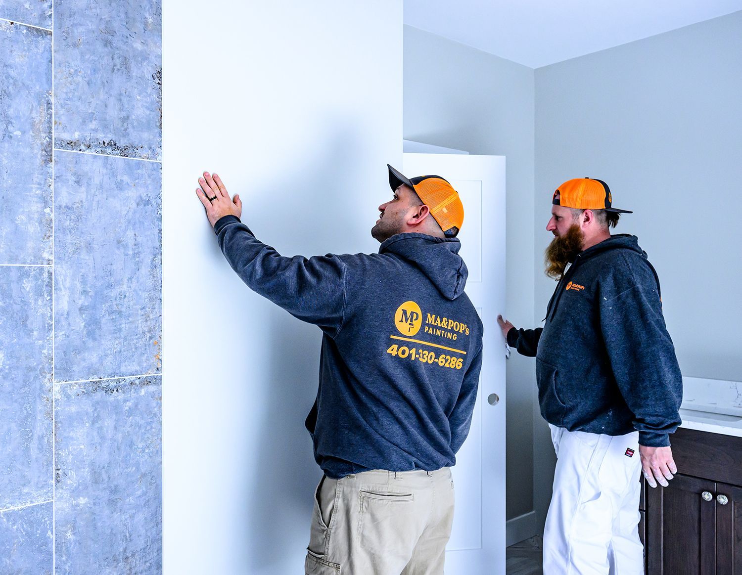 Two painters in matching caps and sweatshirts examine a wall in a room; one touches the wall.