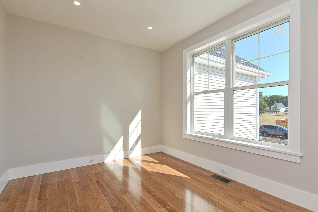 Empty room with hardwood floors, a large window, and off-white walls. Bright sunlight streams in.