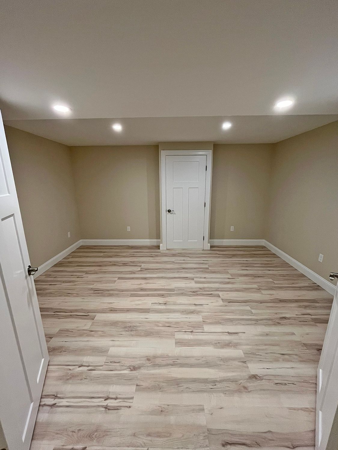 Empty room with light wood-look flooring, beige walls, white ceiling and door, recessed lights.