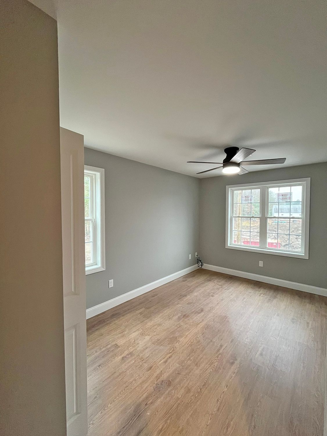 Empty room with wood floors, gray walls, two windows with white trim, and a ceiling fan.