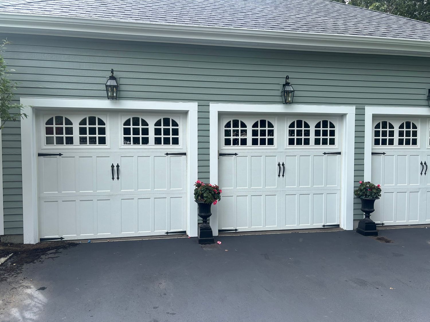 Three white garage doors with arched windows, black hardware, and sconce lights on a teal-sided building.