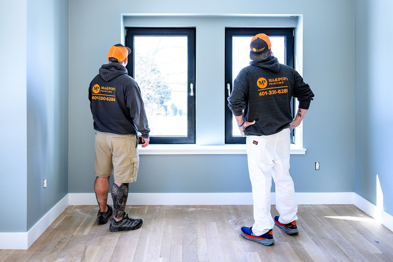 Two workers in hoodies and hats, looking out a window in a freshly painted room.
