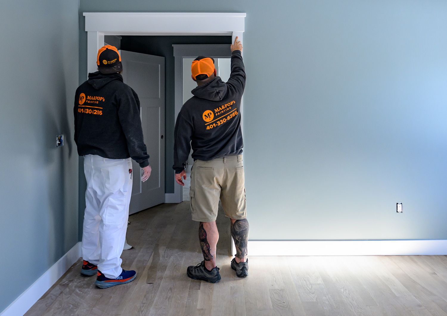 Two workers install white trim around a doorway. One has a prosthetic leg. Interior room, light blue walls.
