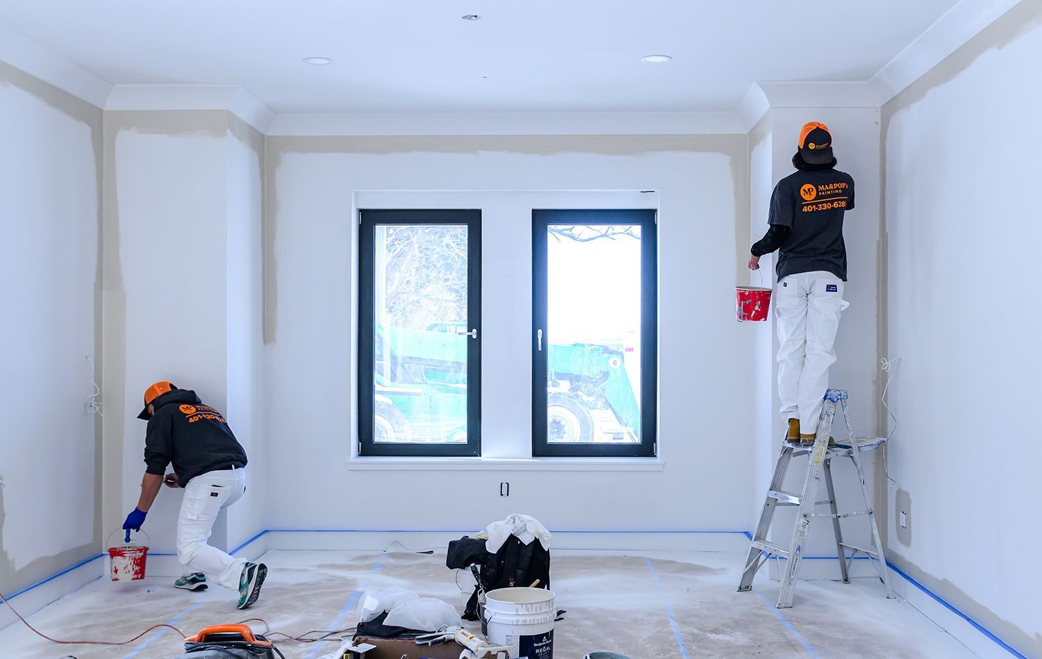 Two painters in orange hats and white coveralls painting a room, one on a ladder, two windows in background.