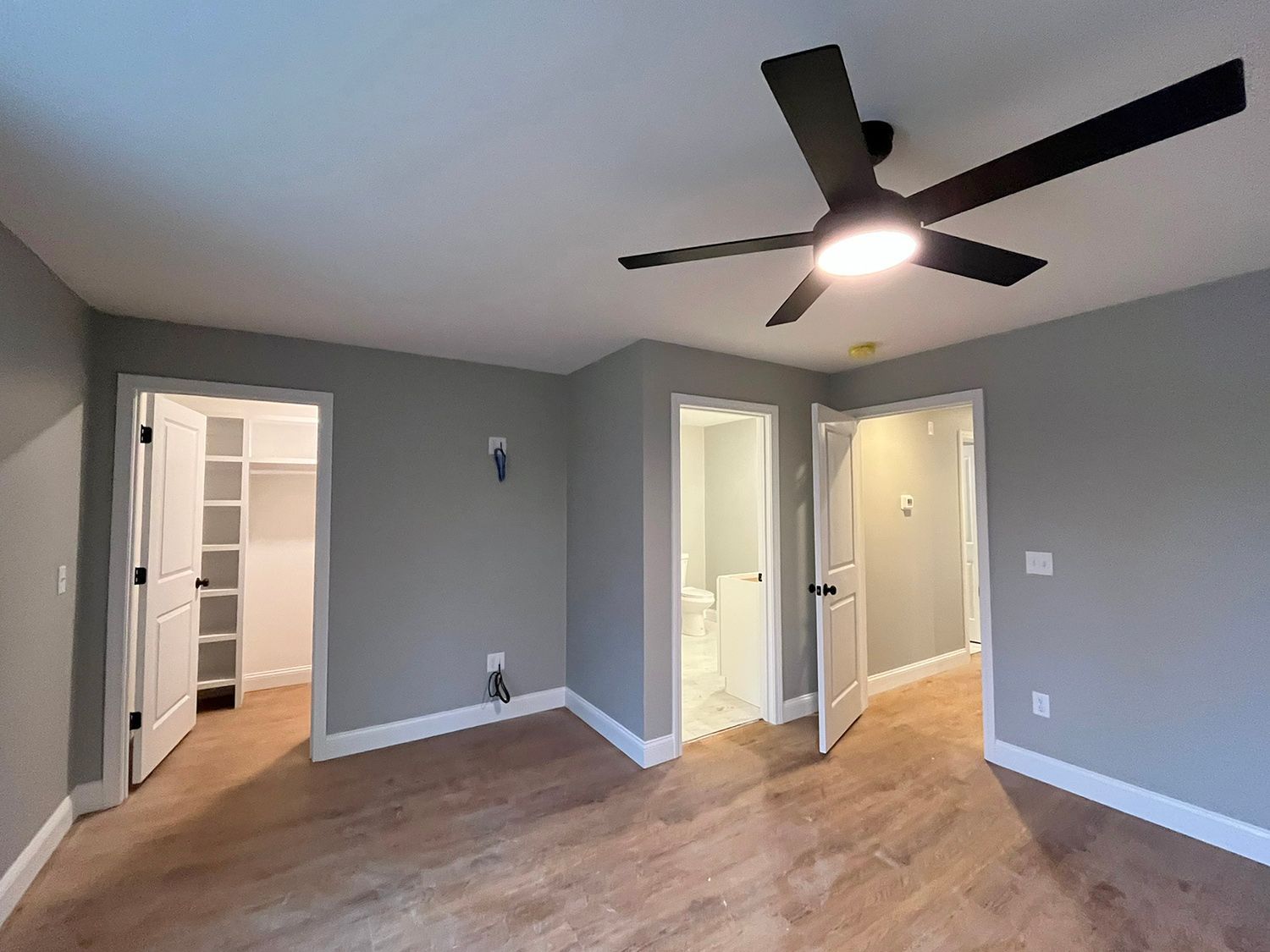 Empty bedroom with open doors to a closet and bathroom. Brown floors, gray walls, black ceiling fan with light.