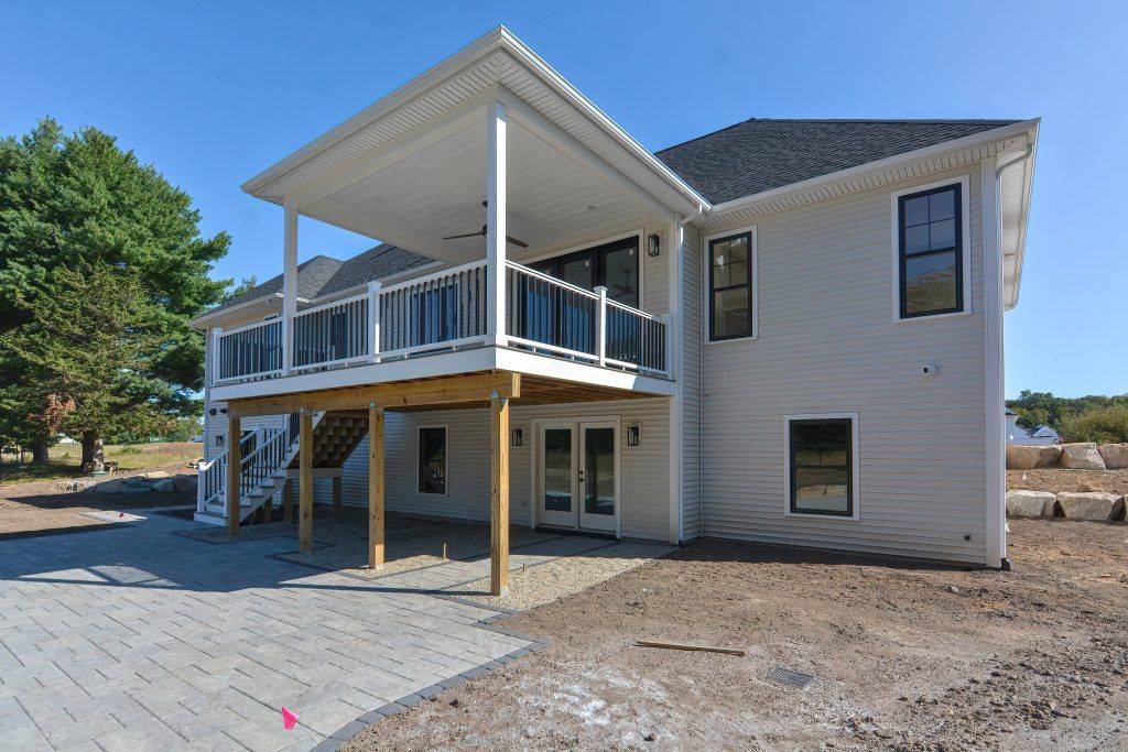 Rear view of a light gray house with a covered deck, black windows, and a paved patio.