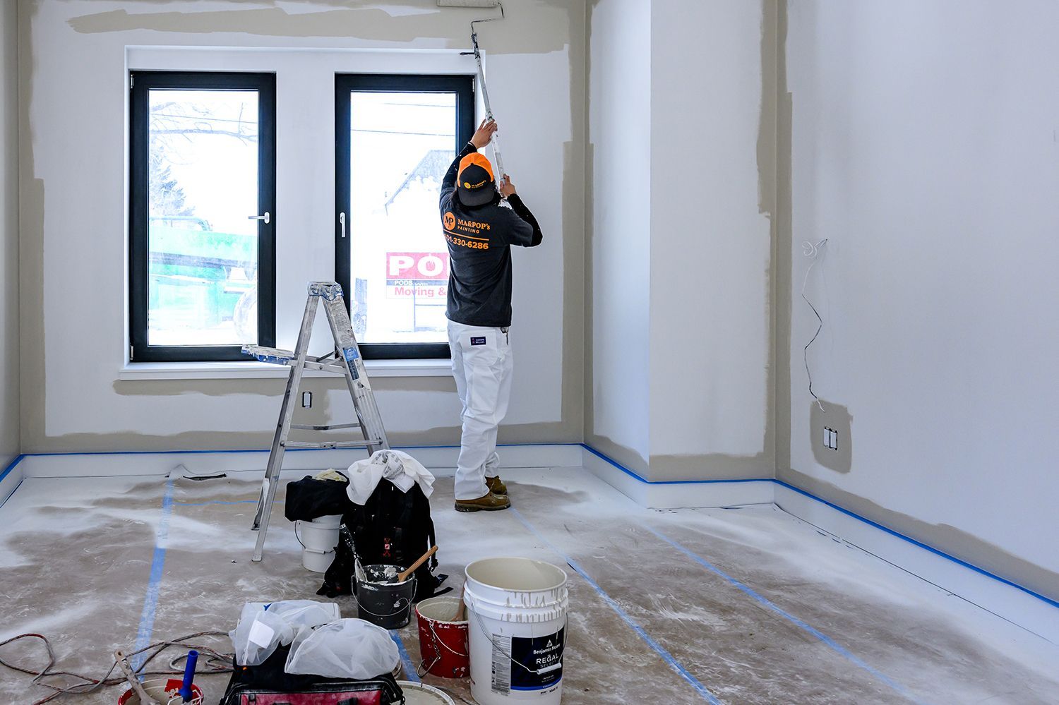 Person painting a room with a roller, standing on a ladder. White walls, blue tape on the floor.